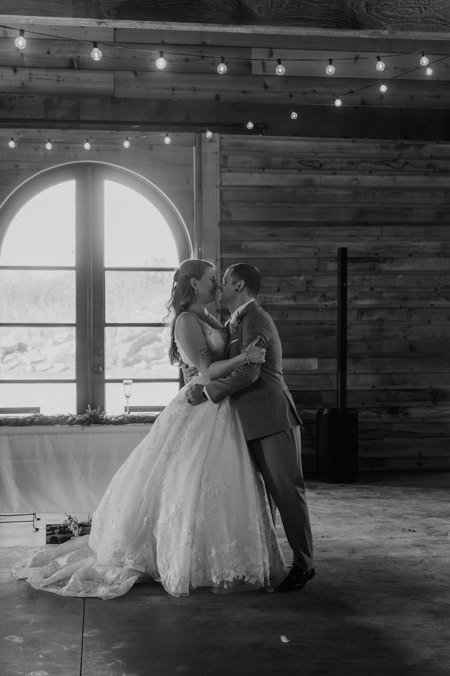 Black and white photo of a bride and groom sharing their first dance kiss in a rustic barn venue with string lights.