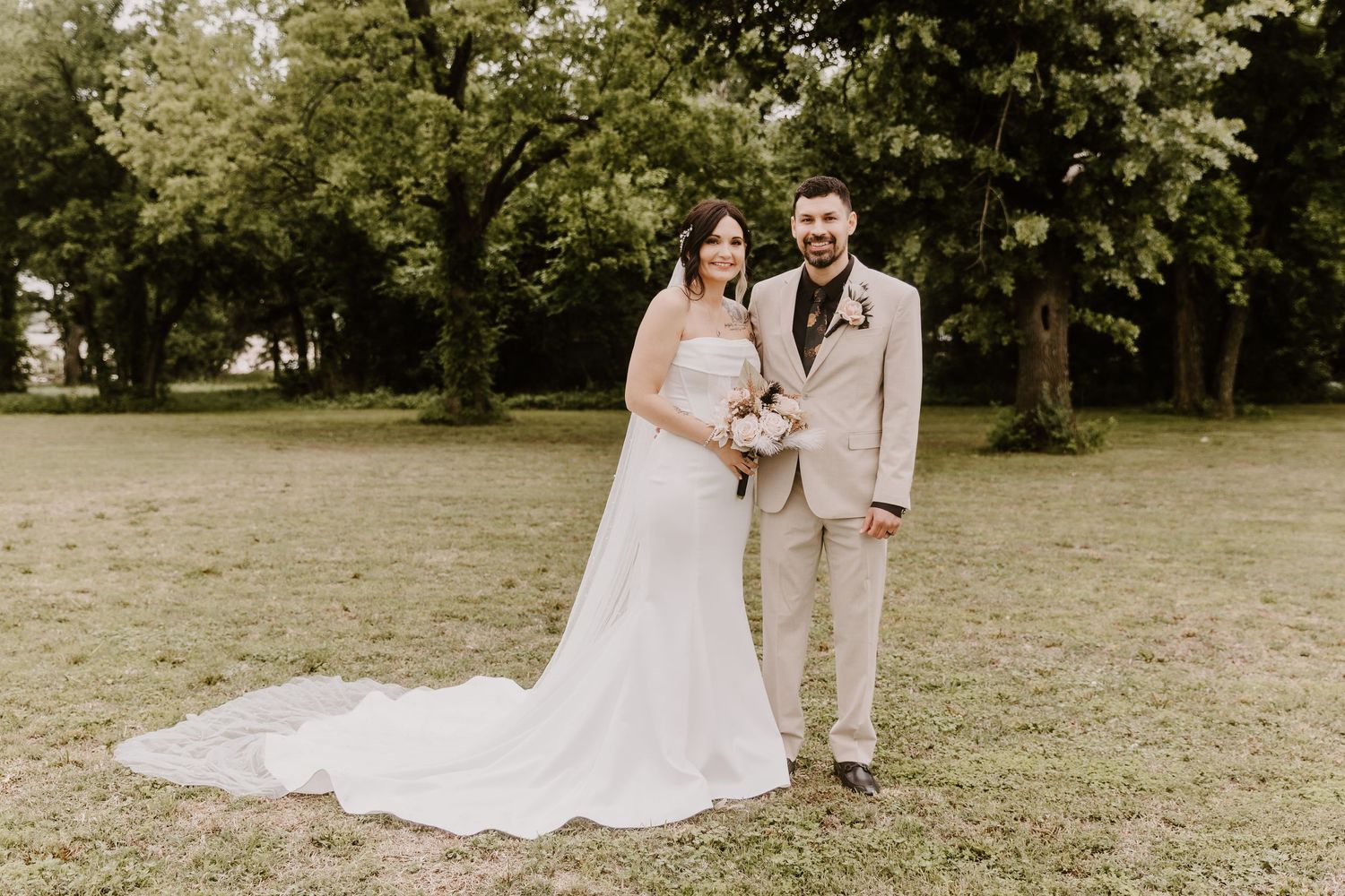 Bride in white gown and groom in tan suit pose together on green lawn surrounded by trees.
