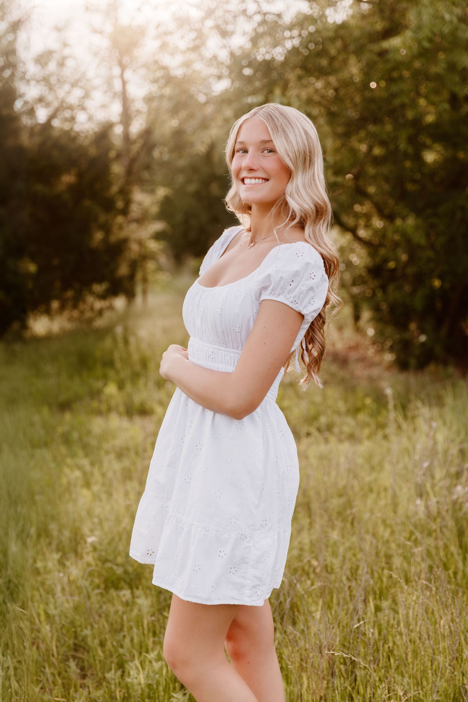 A person in a white summer dress poses outdoors during golden hour in a grassy field with soft lighting.
