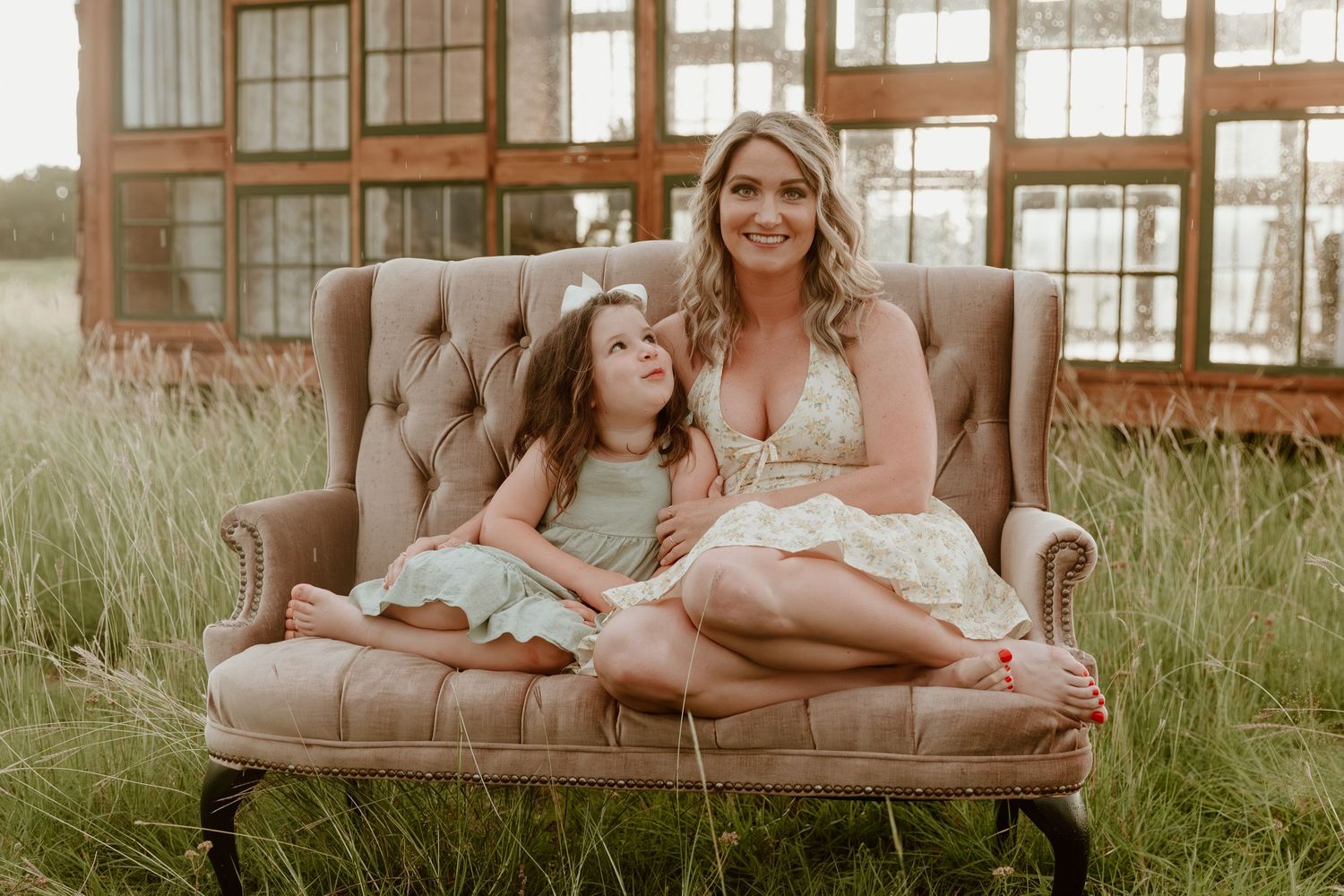 A rustic outdoor setting features a beige velvet couch surrounded by tall grass and wooden fencing at sunset.