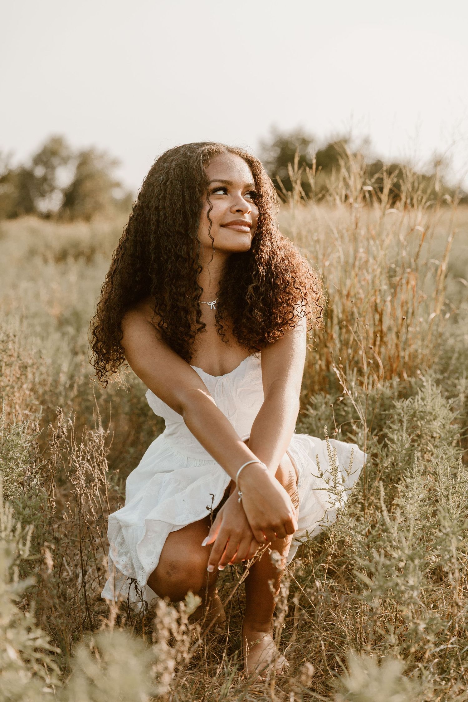 Person in a white sundress poses in a sunlit field of tall grass during golden hour.