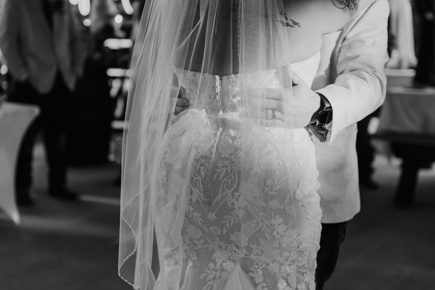 A bride wearing a white lace wedding dress and veil stands in front of a dark background.