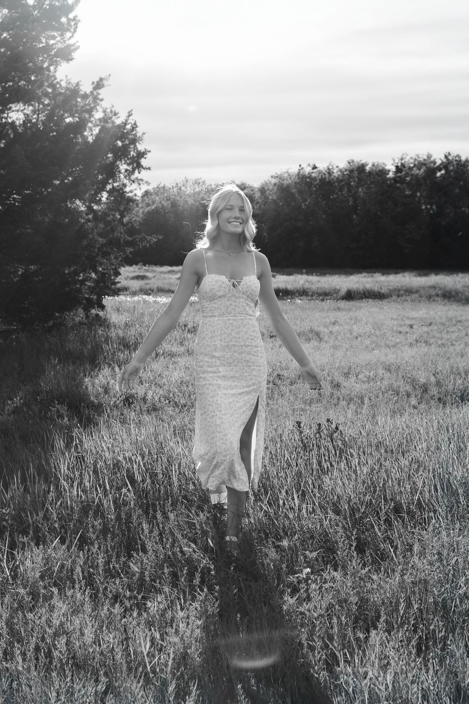 A black and white portrait of someone in a lace dress walking through a sunlit meadow.