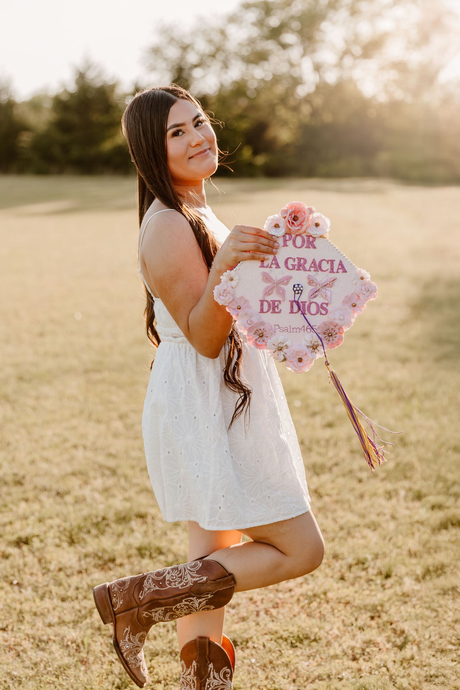 Young woman in white dress and cowboy boots holds pink floral sign reading 'La Gracia De Dios' in sunlit field.