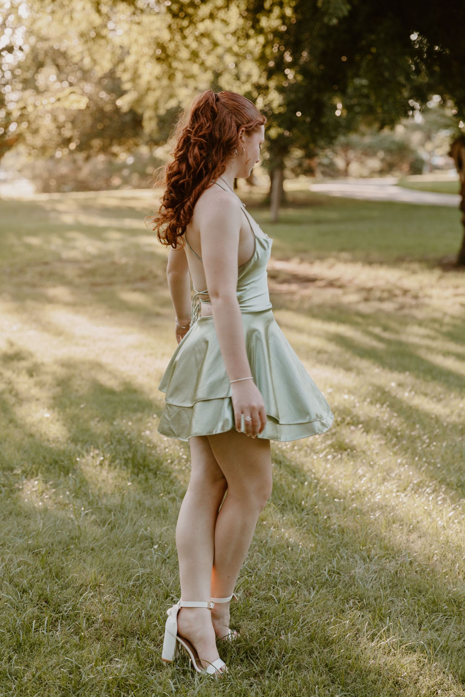 A person in a mint green dress with flowing red hair poses in golden sunlight on grass wearing white heels.