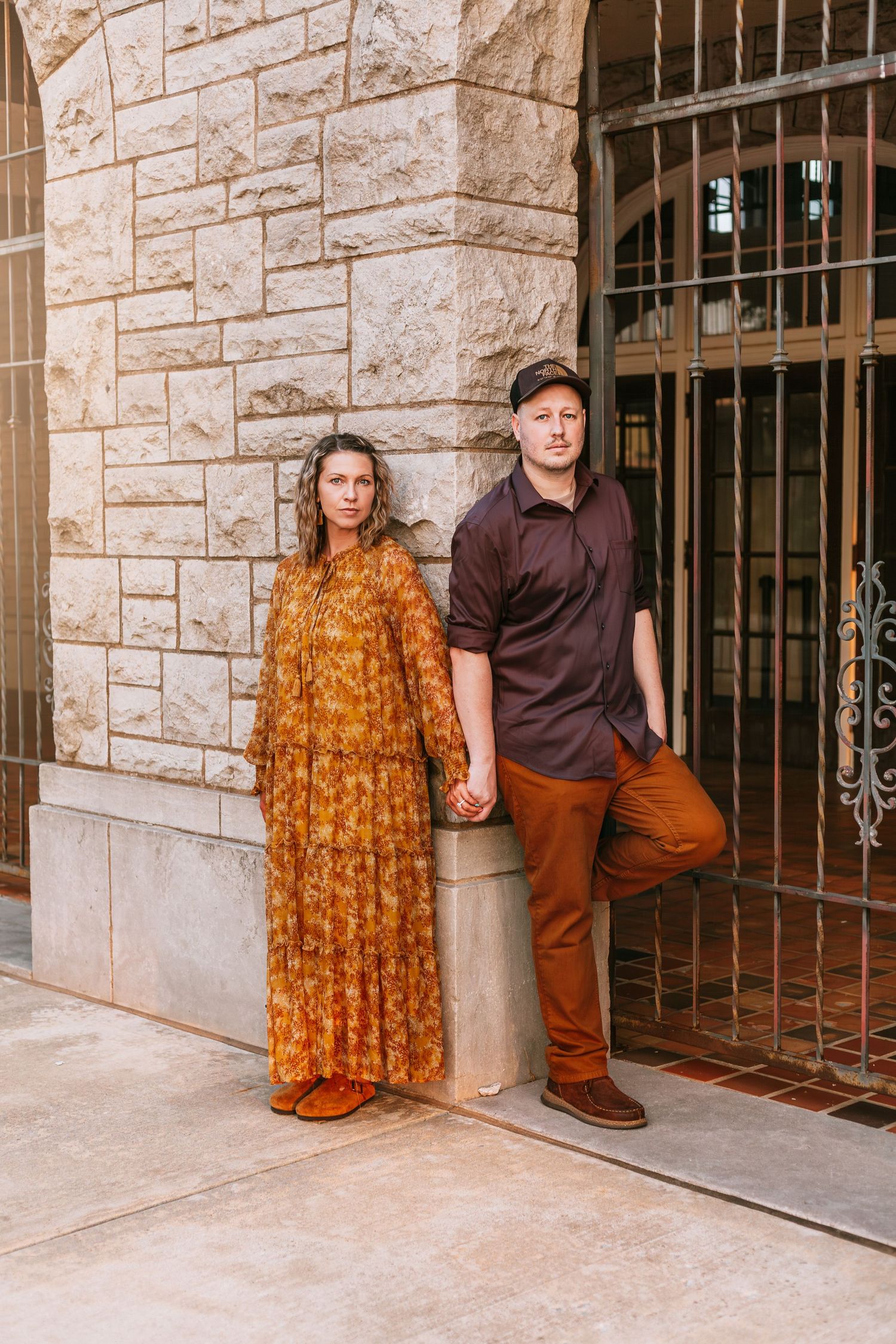 Couple in casual attire poses by stone building with ornate metal gates.