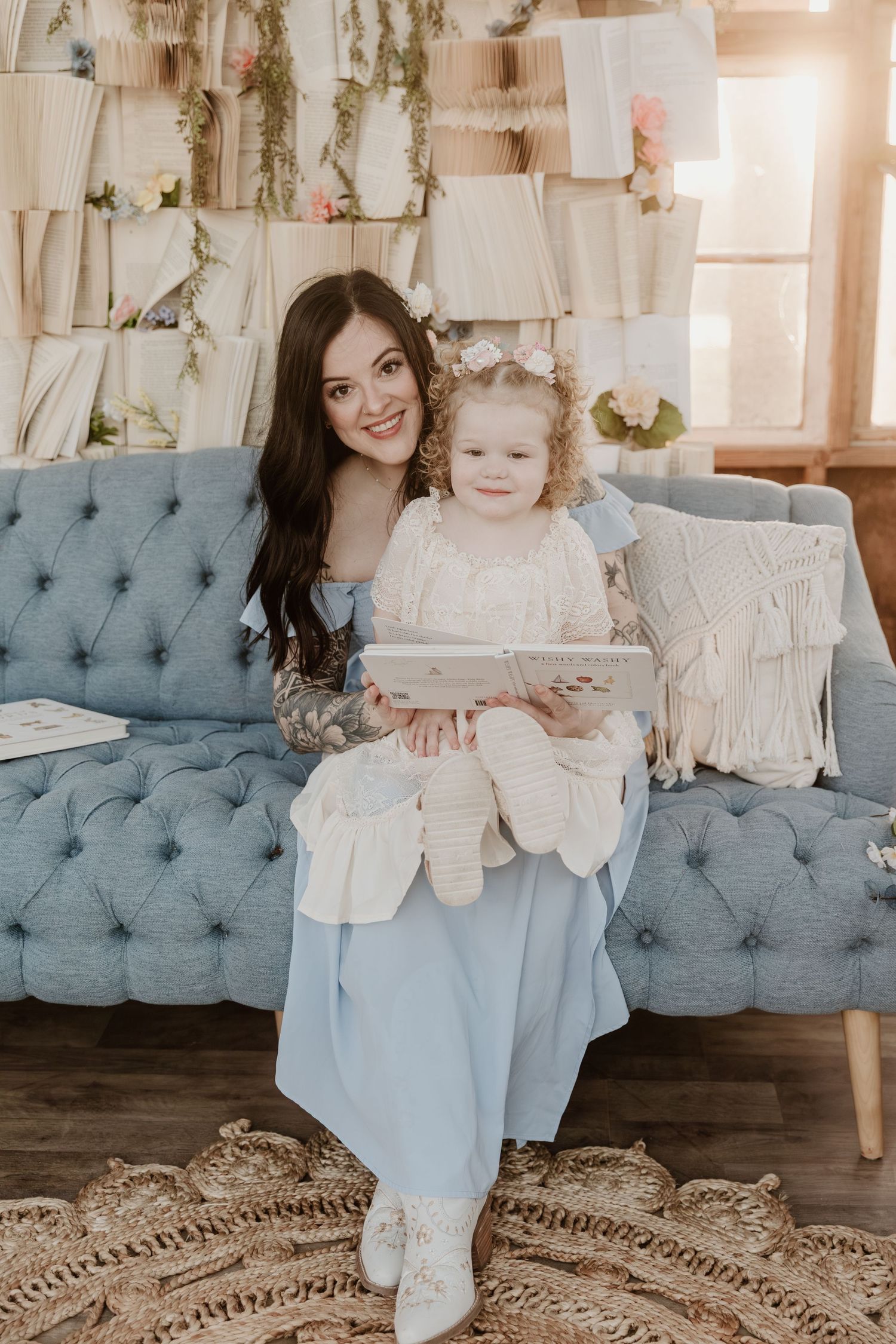 A smiling mother in a blue dress holds her toddler daughter on a tufted blue sofa, reading a book together.