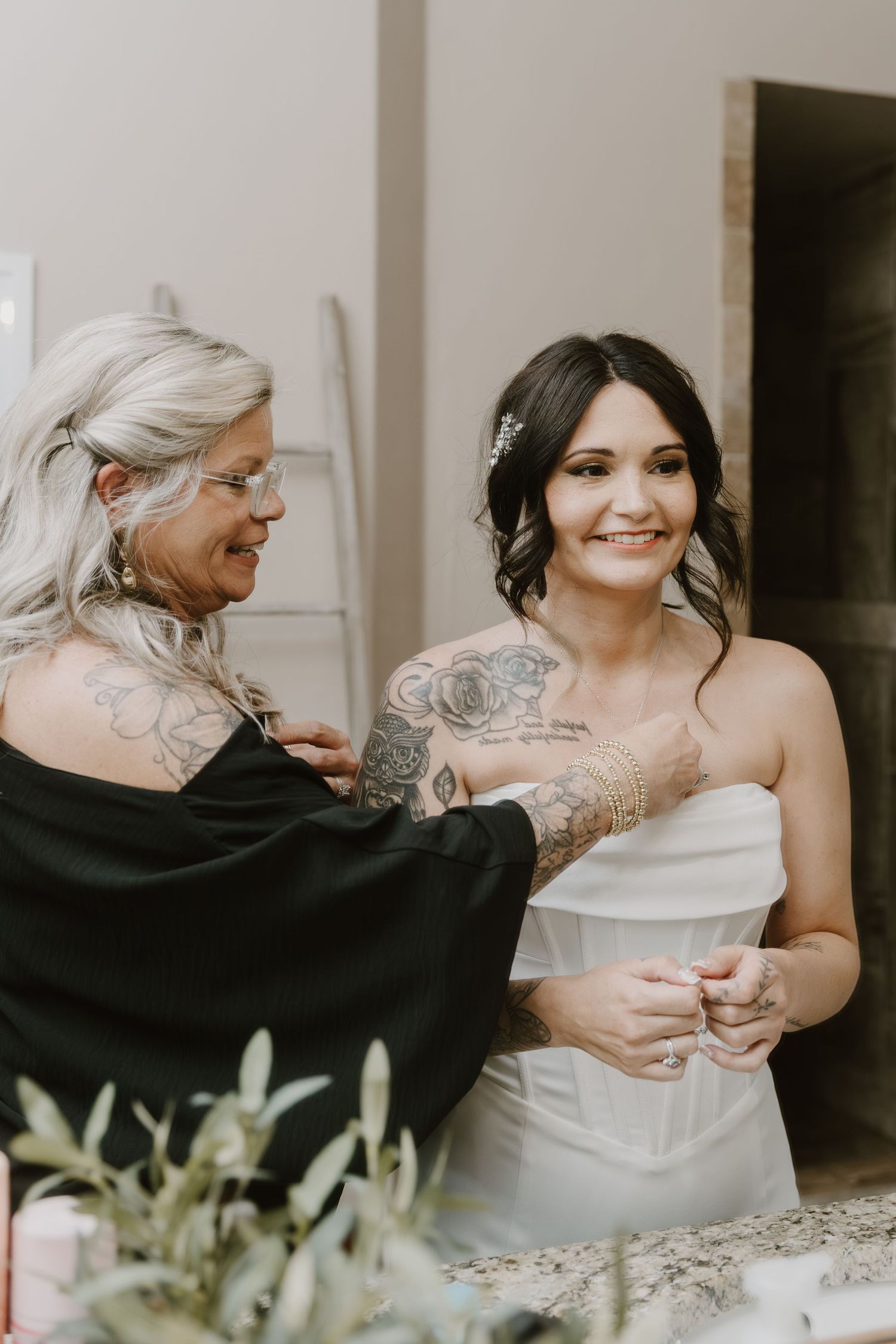 A bride in a white strapless gown smiles as a tattooed woman with gray hair helps her prepare for her wedding day.