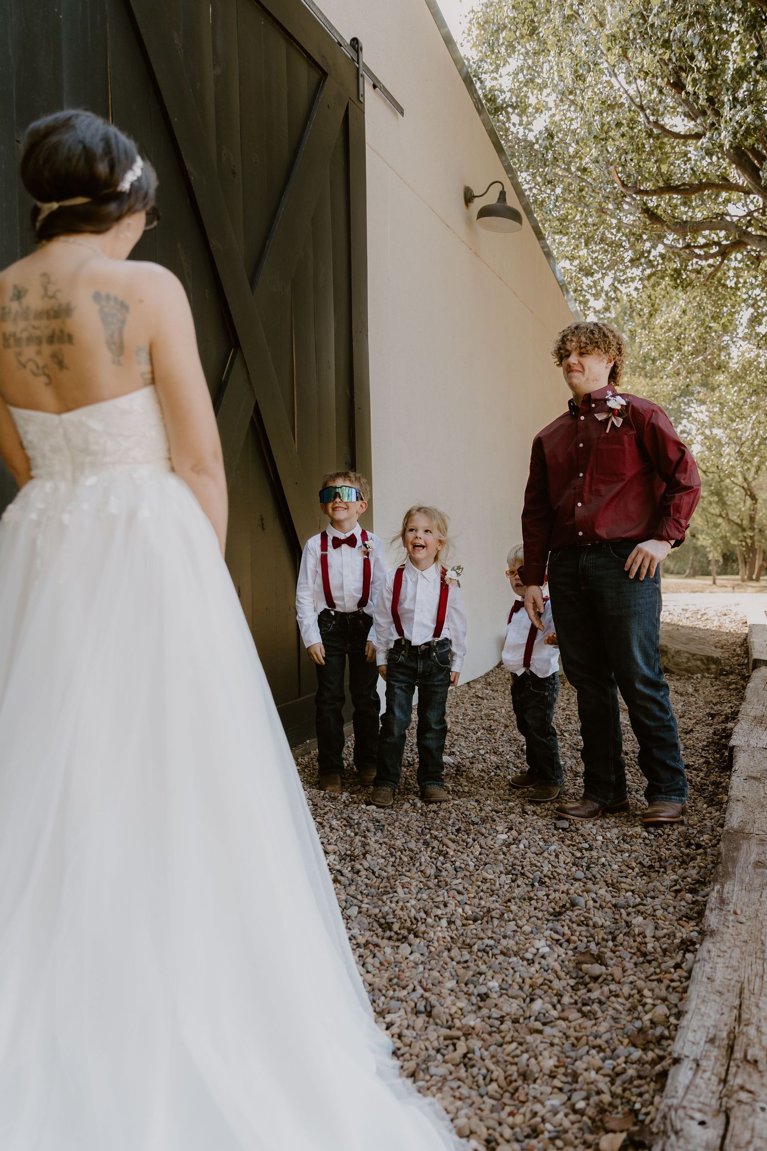 Wedding party in burgundy and white attire gather outside rustic barn doors for candid moments.