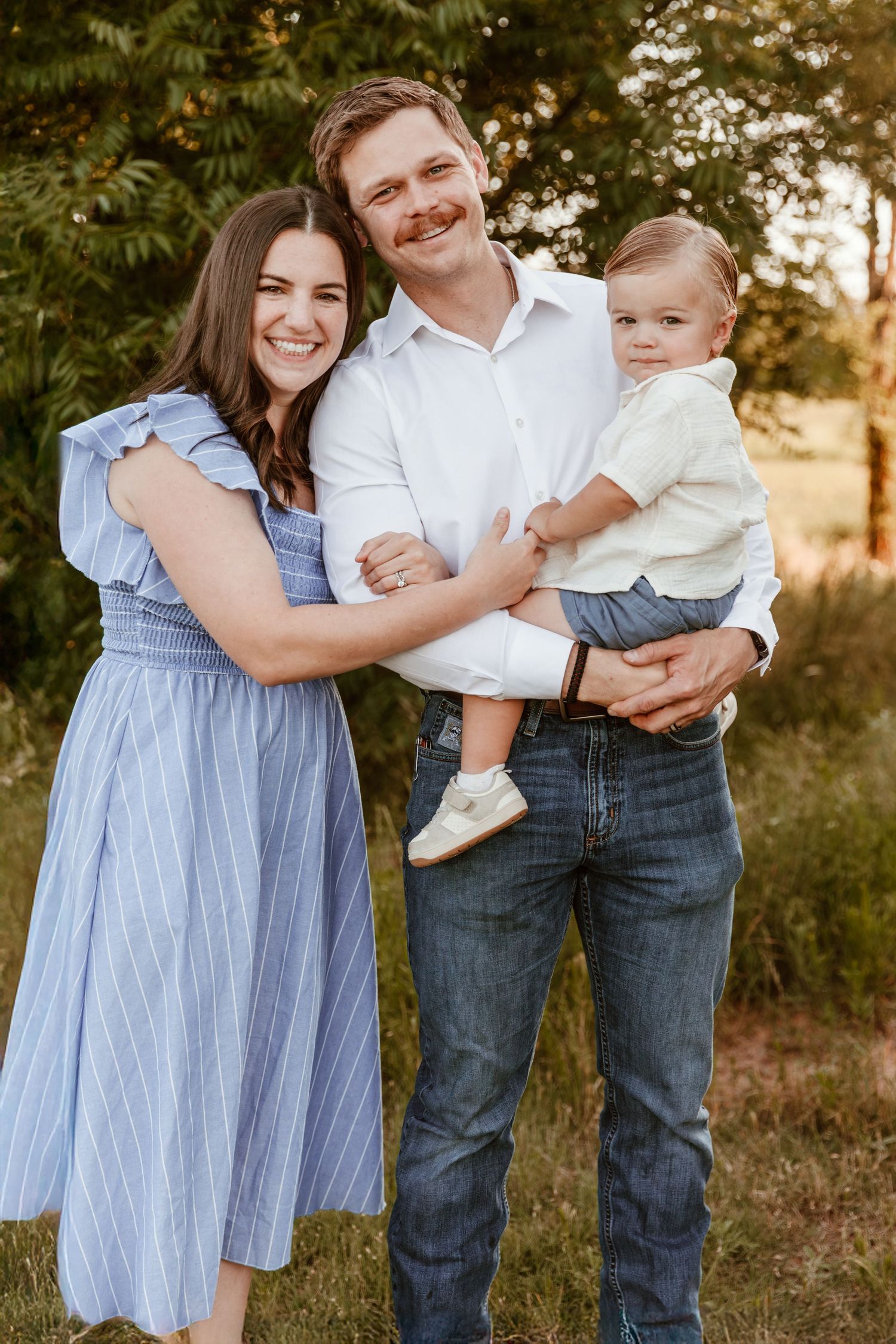 A family photoshoot shows joyful interactions between parents and a baby in a natural outdoor setting at sunset.