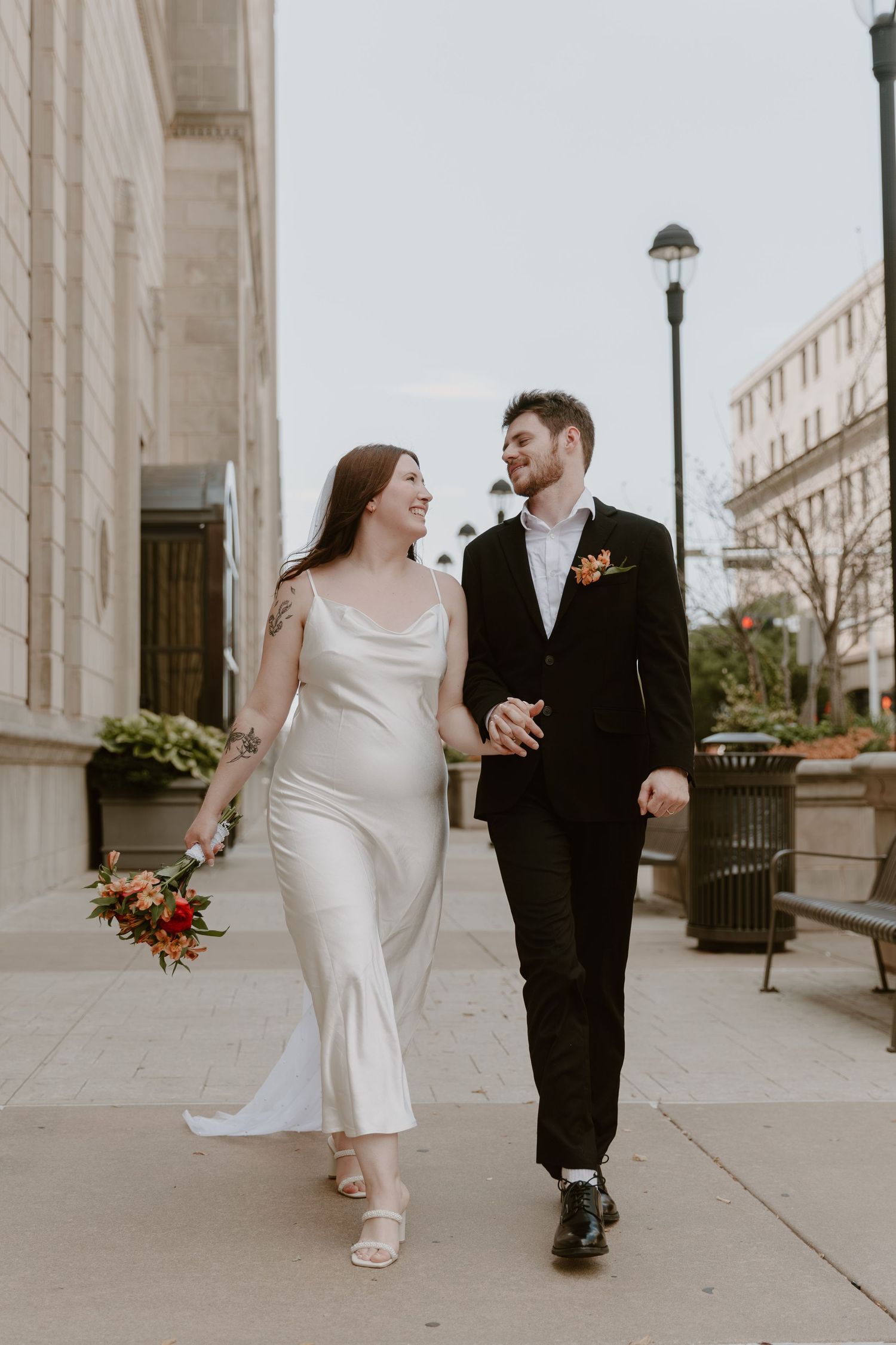 A couple in formal wedding attire shares candid laughing moments walking along a grand building's exterior columns.