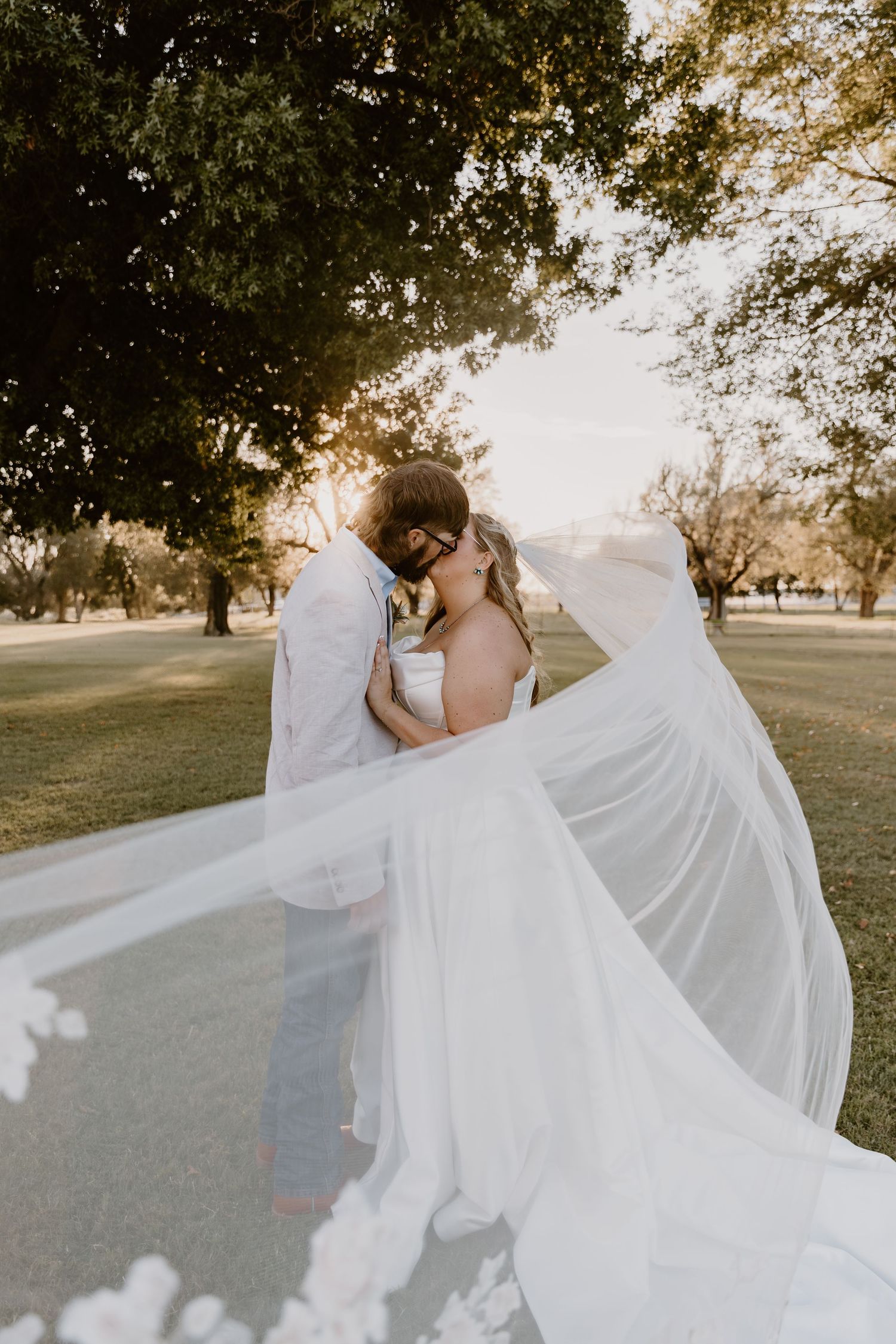 A flowing white wedding dress veil billows dramatically in the breeze during a golden hour outdoor photoshoot.
