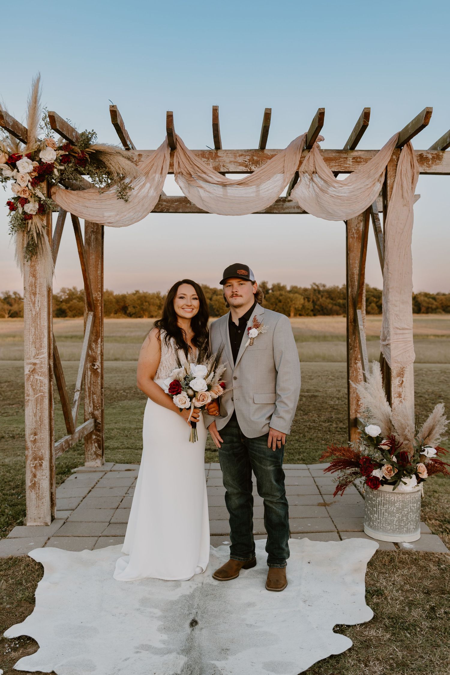 A couple poses together under a rustic wooden pergola decorated with flowing white fabric and floral arrangements.