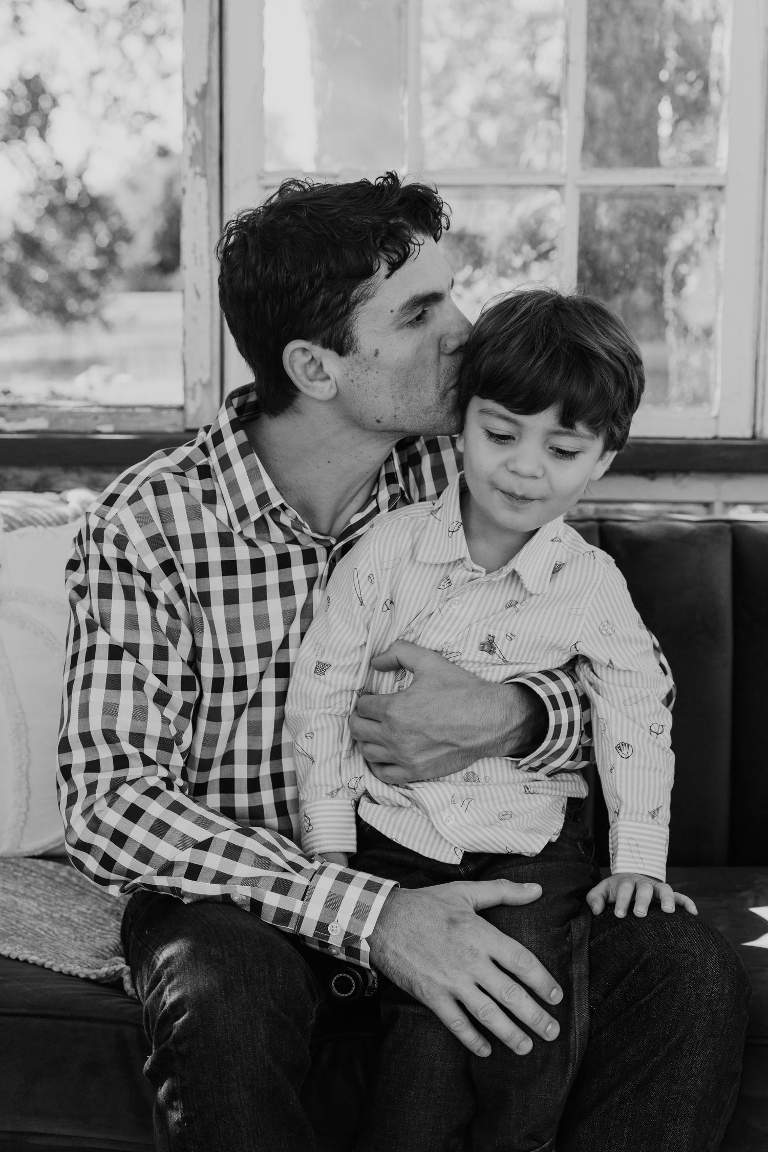 A tender black and white portrait of a parent and child sharing an affectionate moment on a bench by a window.