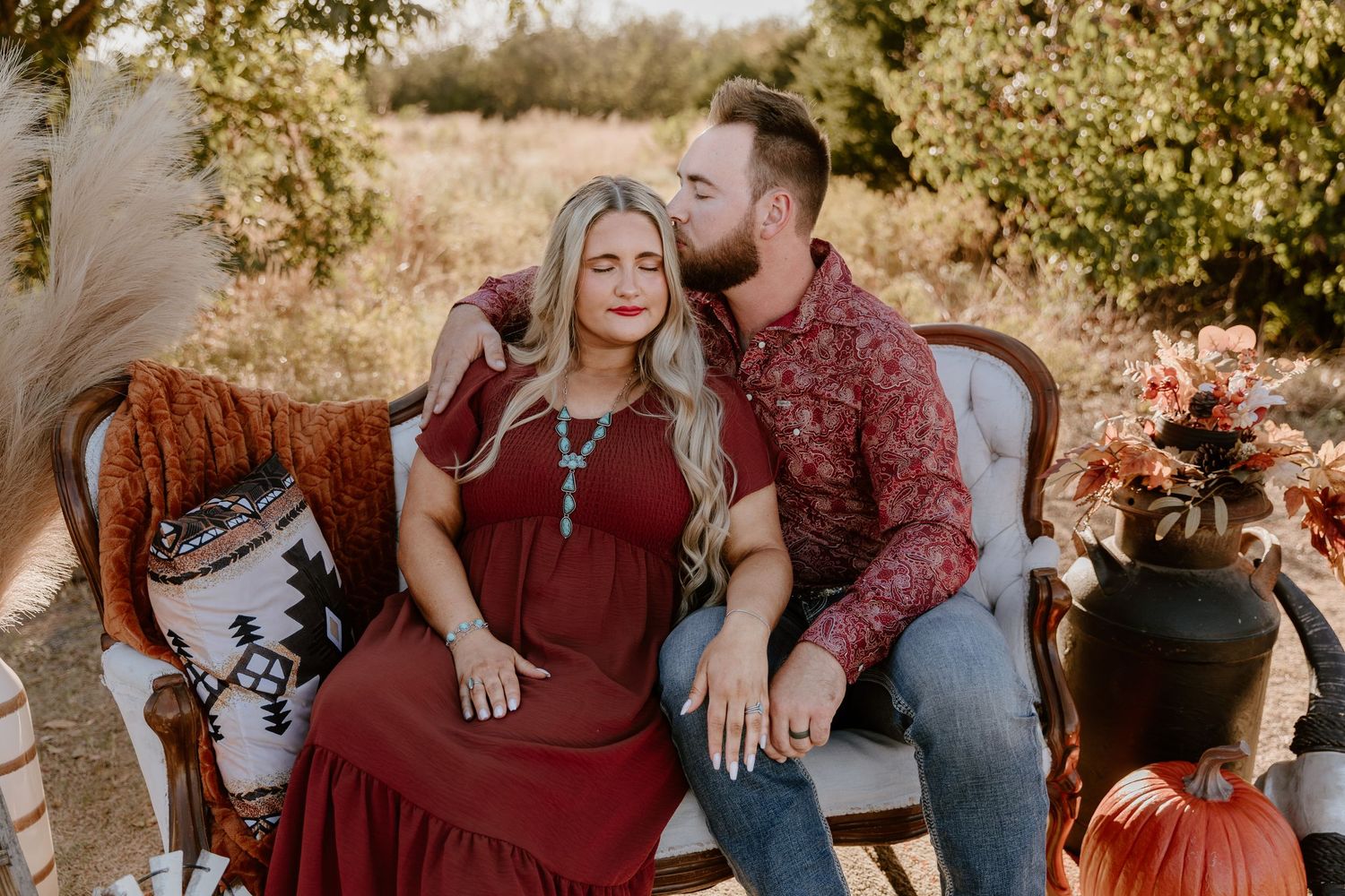 Couple sits on rustic outdoor furniture surrounded by autumn decor with pumpkins and dried flowers.