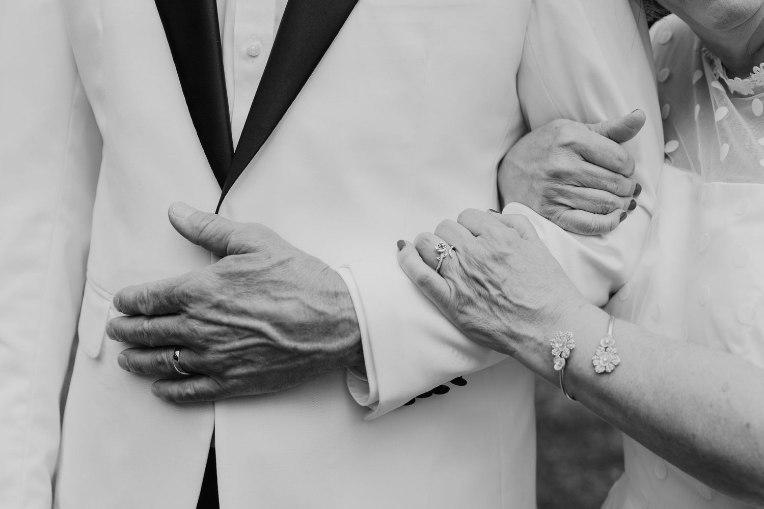 Black and white photo showing hands holding arms while wearing formal attire in an intimate gesture.