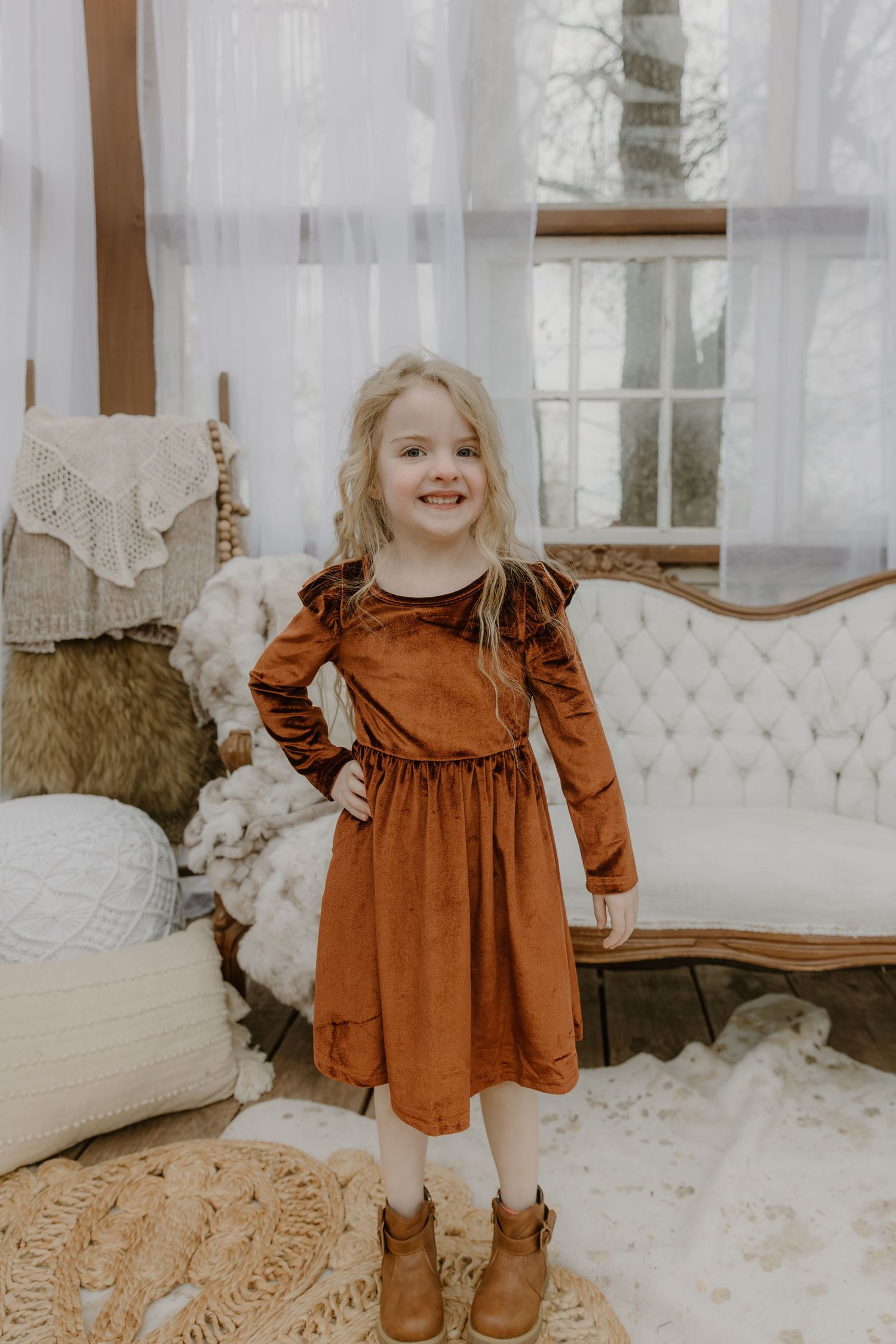 A child in a rust-colored corduroy dress stands in a bright, airy room with white curtains and vintage furniture.
