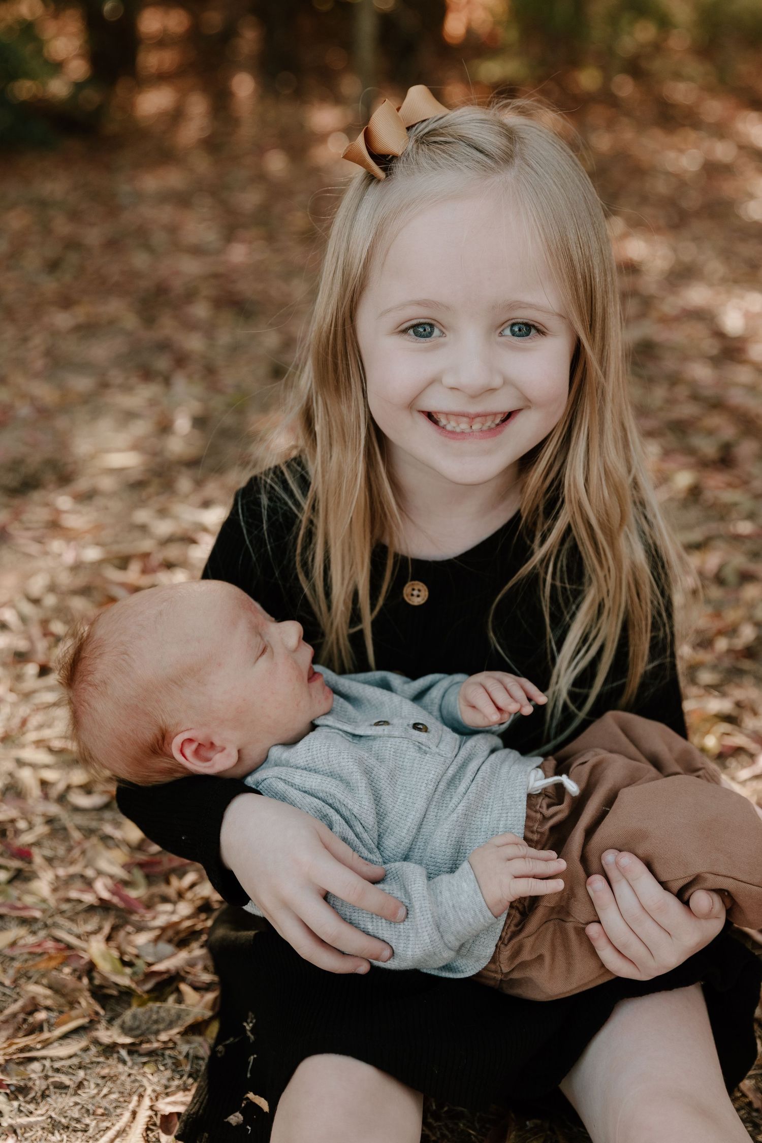 A child in a black dress holds and smiles at a baby wearing grey clothing while sitting on autumn leaves.