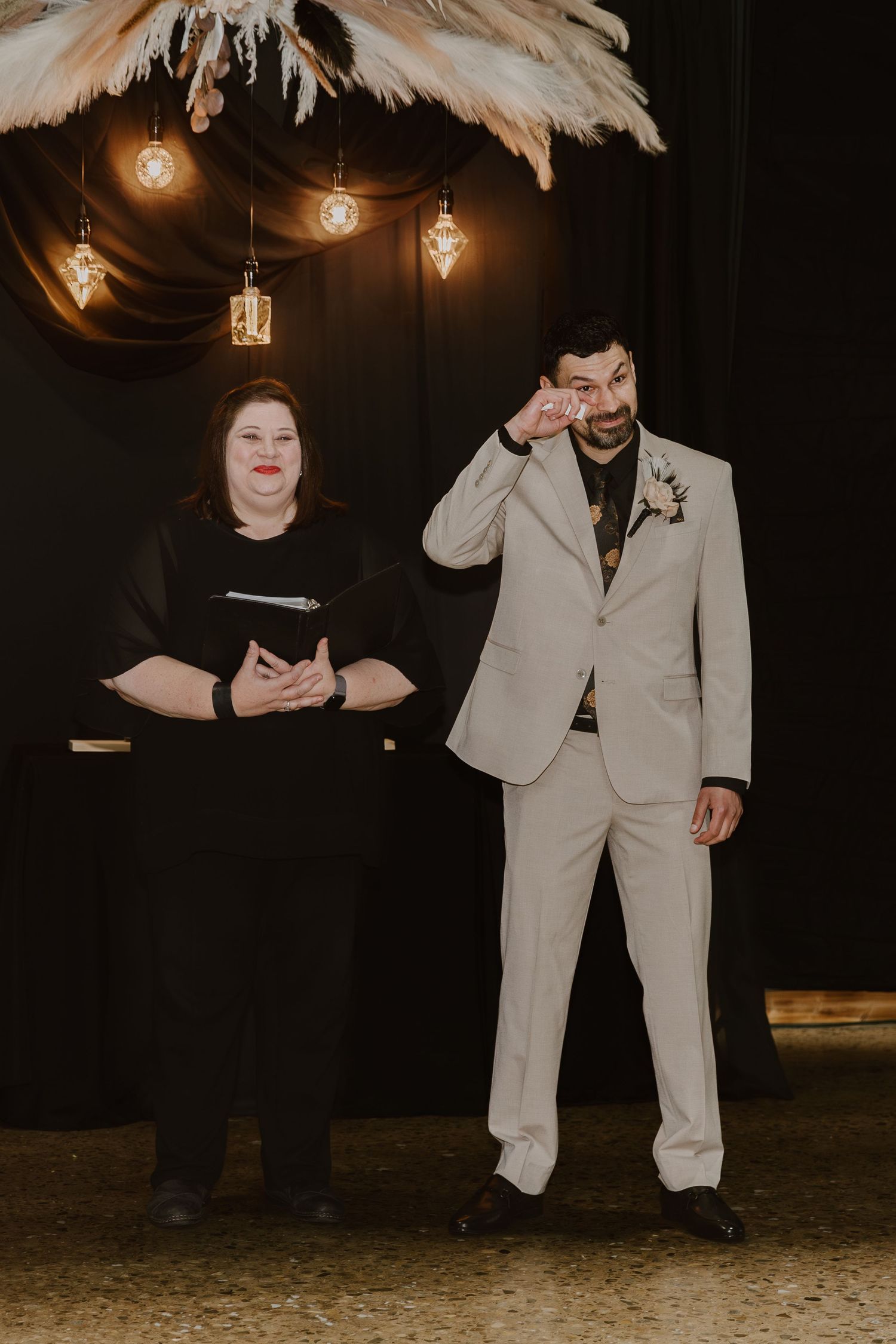 Groom in beige suit wiping tears beside officiant in black at wedding ceremony with pampas grass decor.
