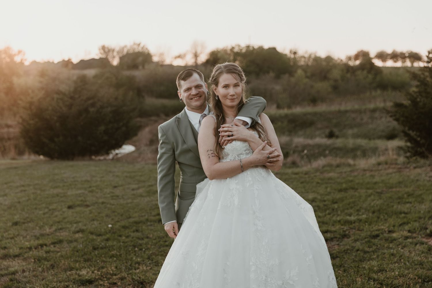 A bride and groom embrace at golden hour in a green field, she in a white ball gown, he in a gray suit.