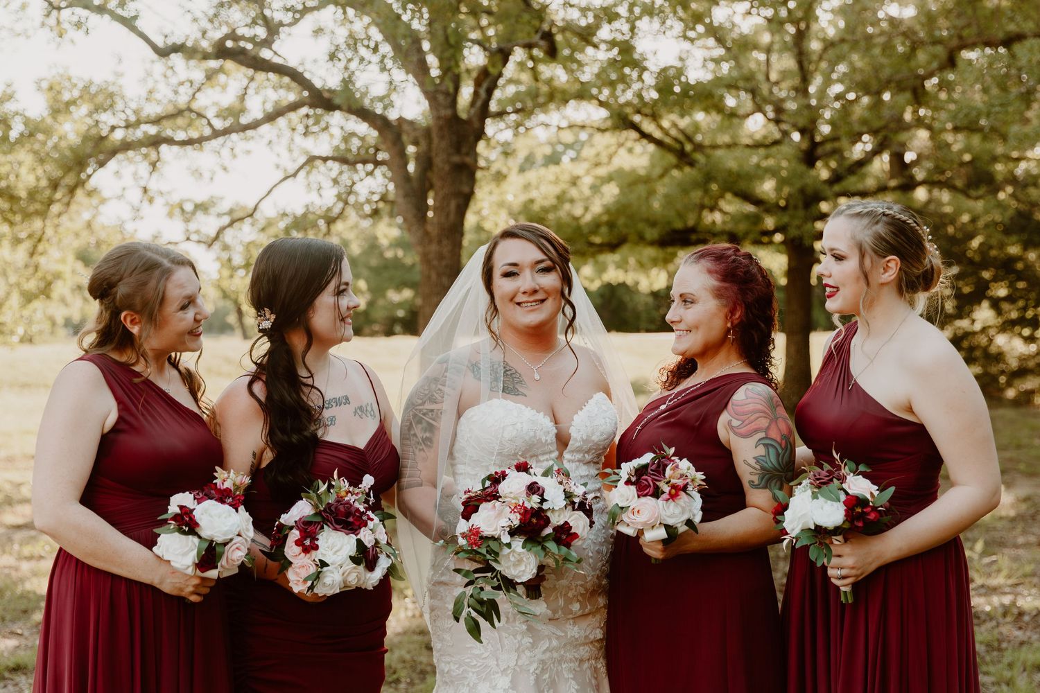 Wedding party in burgundy bridesmaid dresses share laughter while holding white and red floral bouquets in outdoor setting.