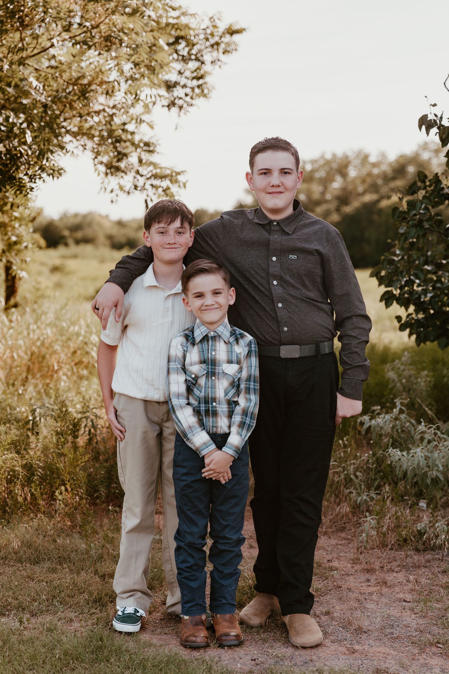 Three people pose together outdoors in a natural setting during golden hour on a dirt path among trees and foliage.