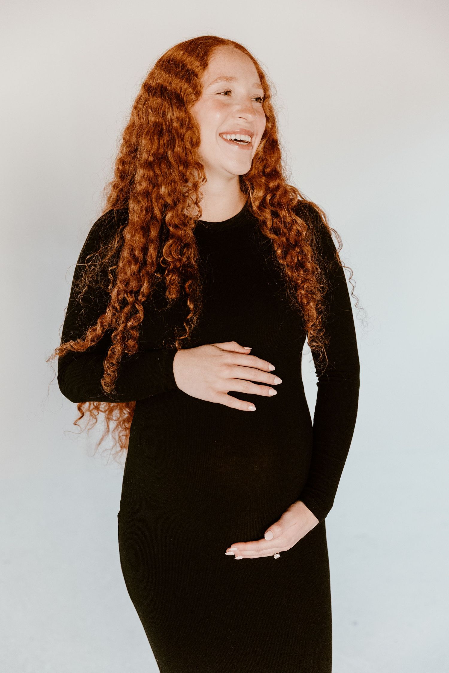 Person with long curly red hair wearing black sweater laughs joyfully against white background in portrait series.