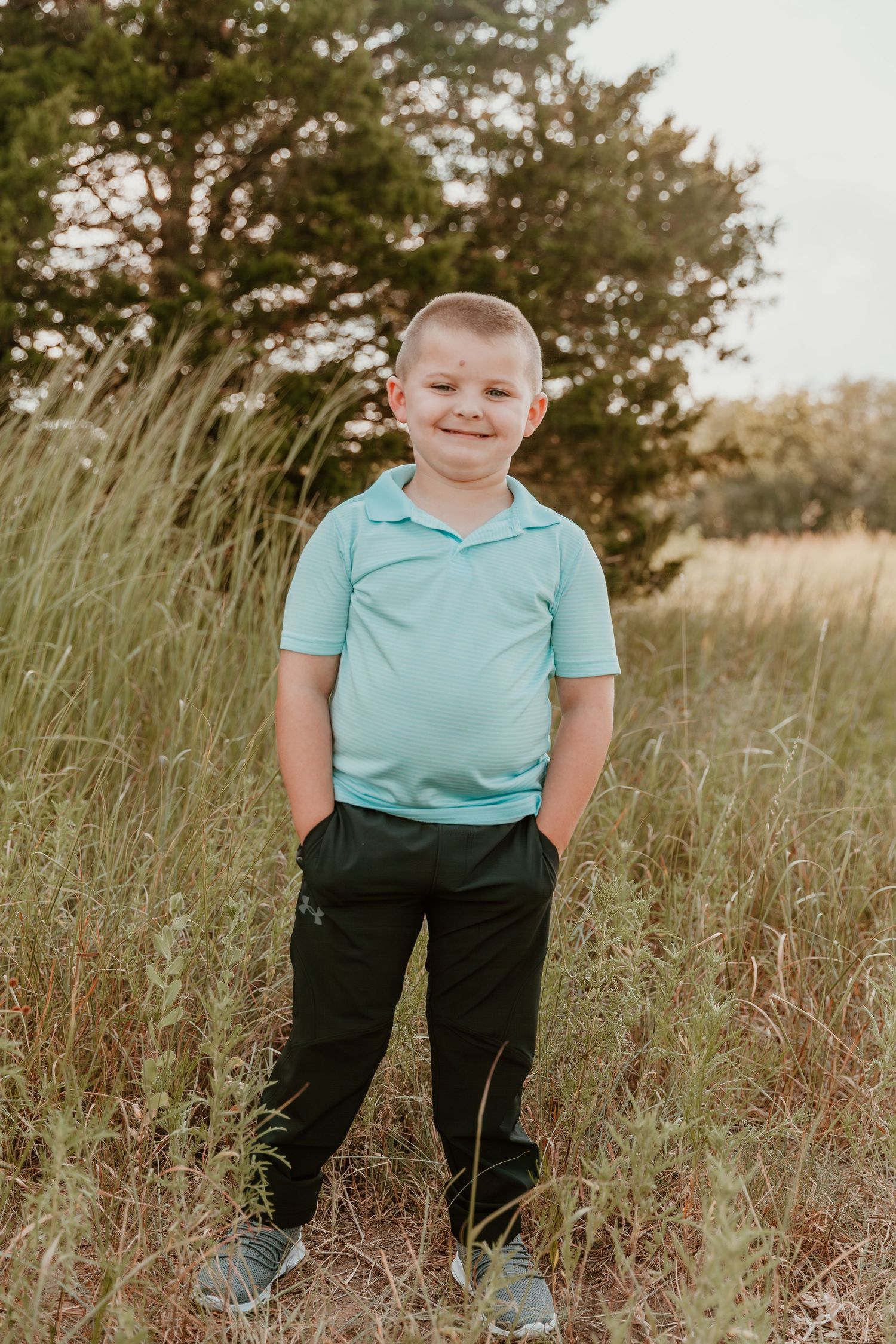 A child wearing a turquoise polo shirt and dark pants stands in a grassy field during golden hour.