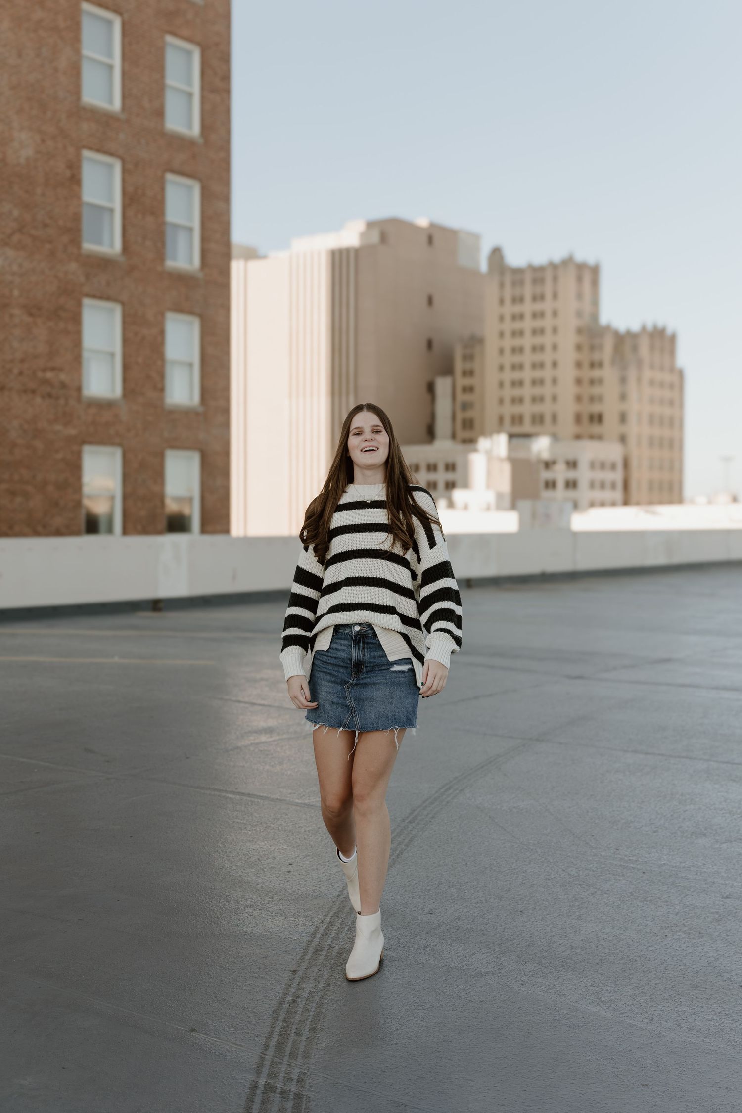 A person wearing a striped sweater and denim shorts walks across an urban rooftop with city buildings in the background.