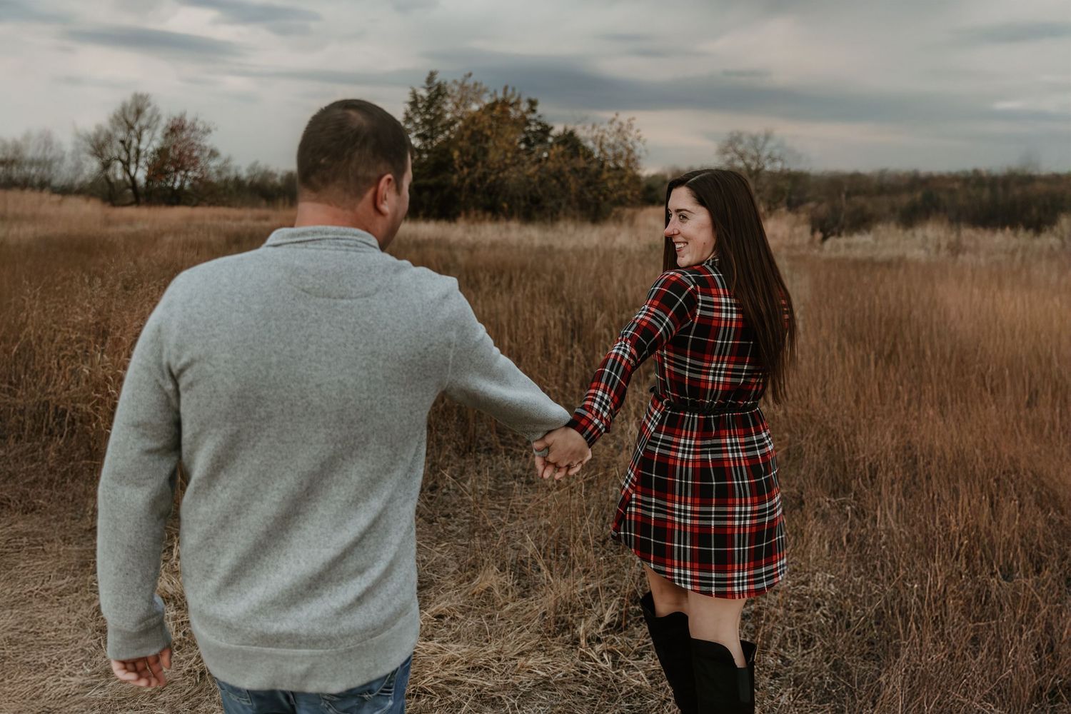 A couple stands together in a golden autumn field during sunset with dramatic cloudy skies overhead.