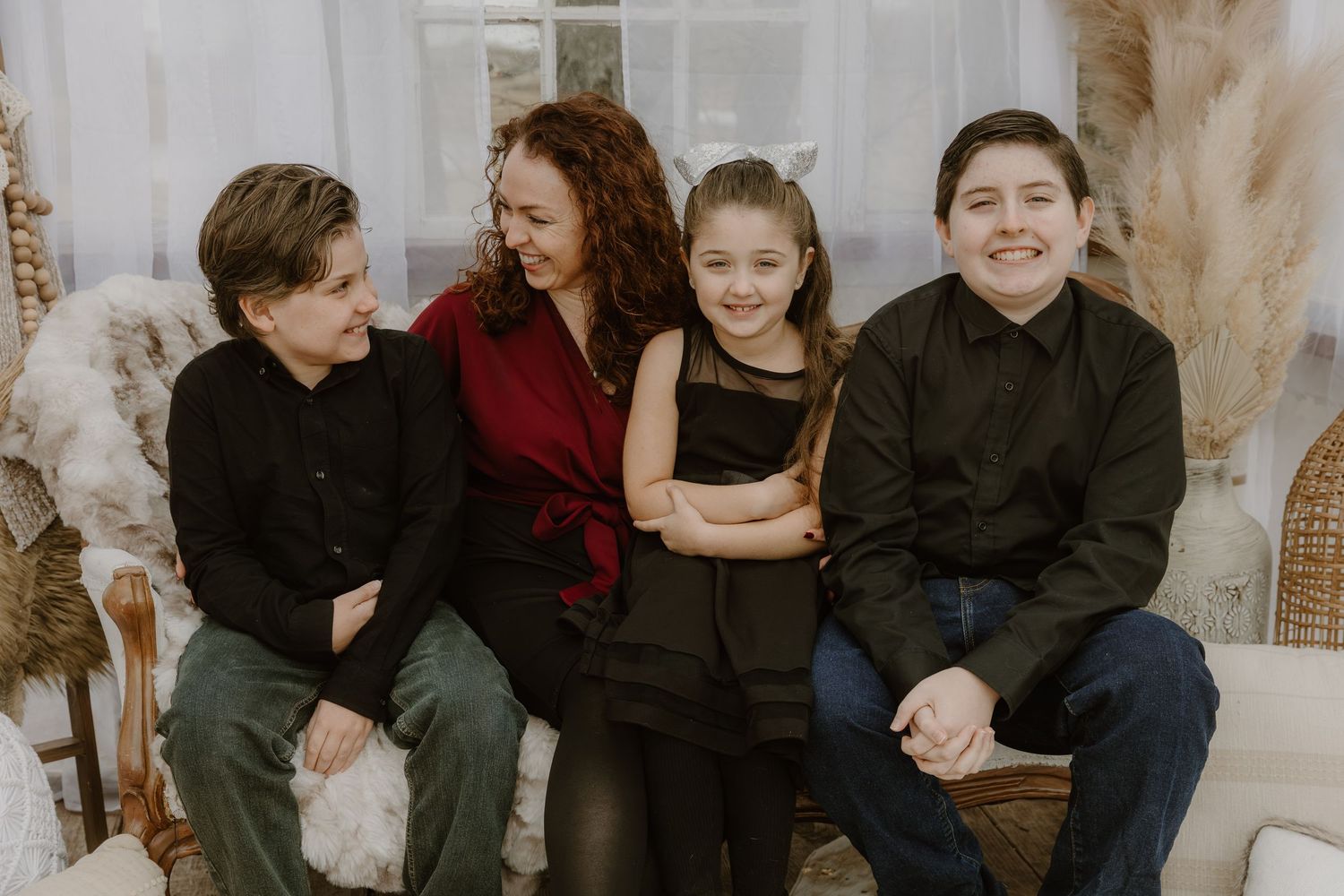 A family of four sits together on a couch, wearing dark clothing against a light neutral background with dried pampas grass.