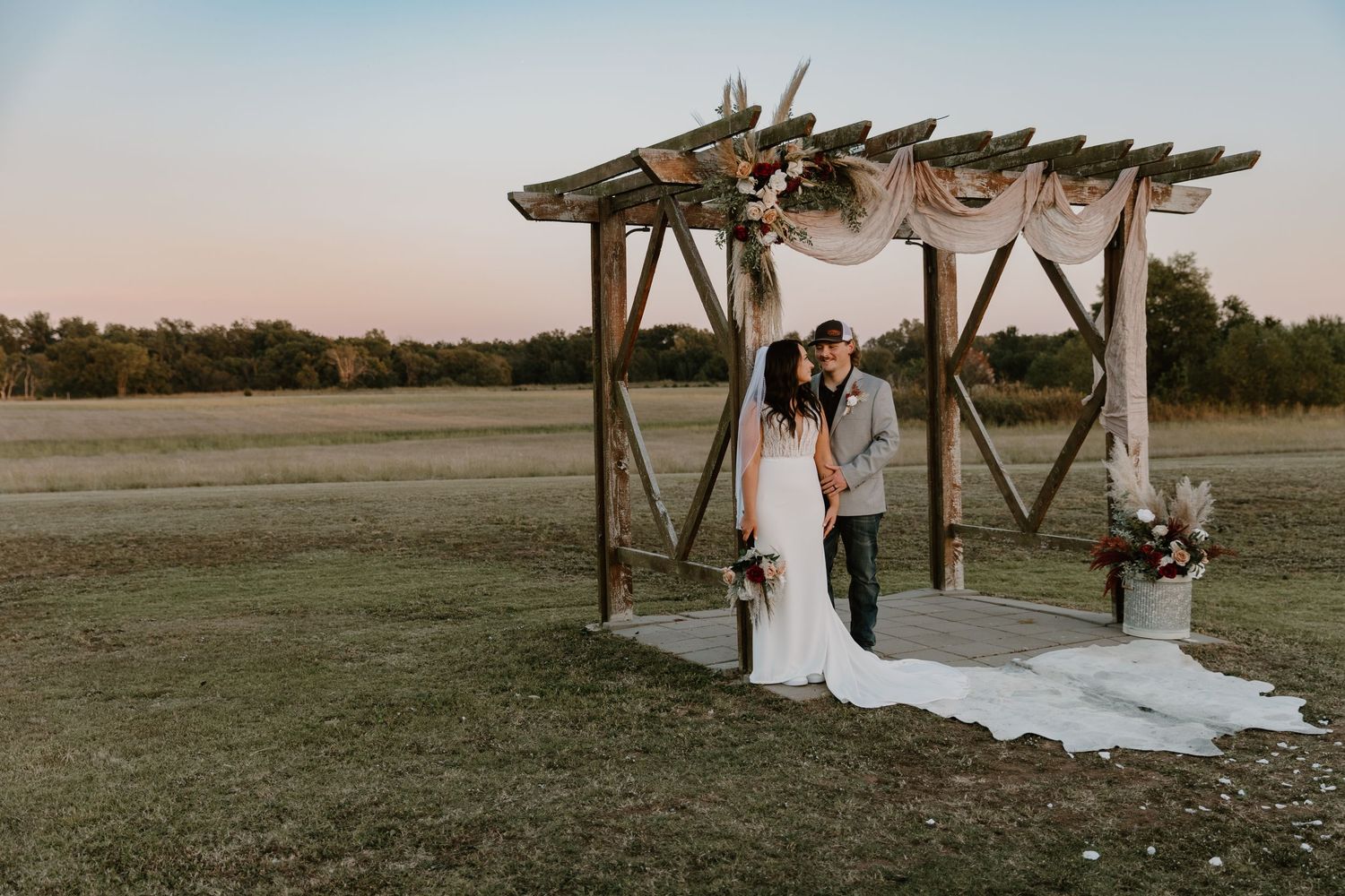 A wedding ceremony takes place under a rustic wooden arbor in an open field at sunset.