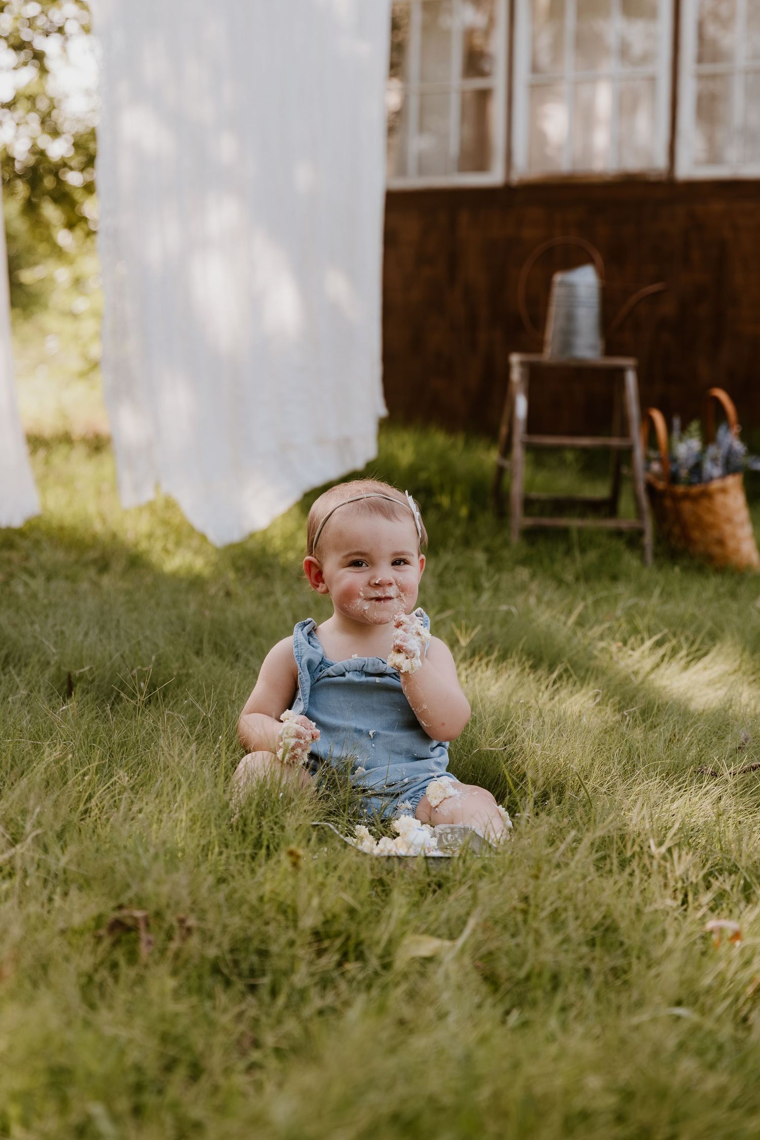 A happy baby girl in a blue romper sits in green grass outdoors, holding a flower in a sunny garden setting.
