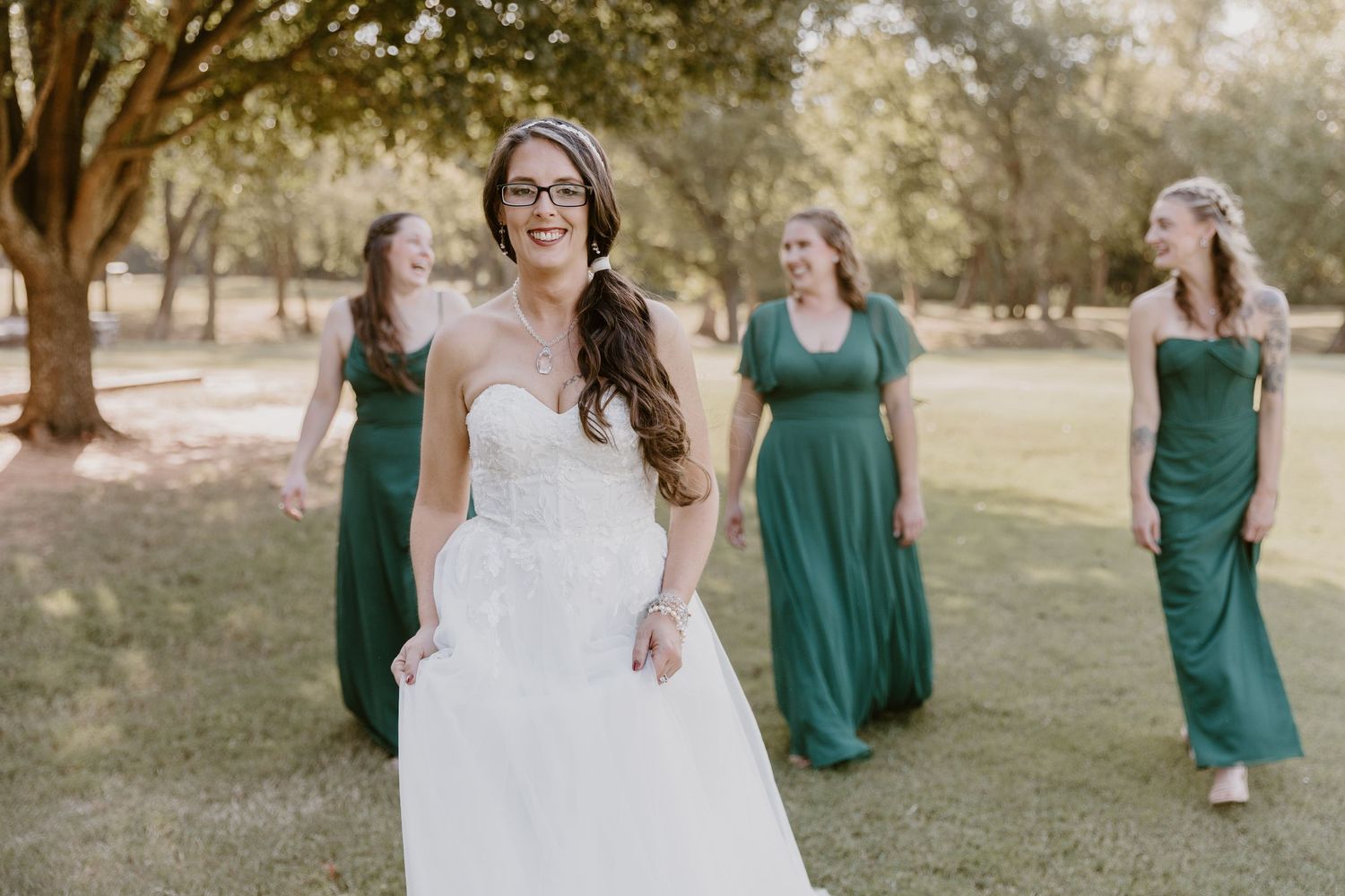 A group of bridesmaids in emerald green dresses walk together in a sunny outdoor setting.