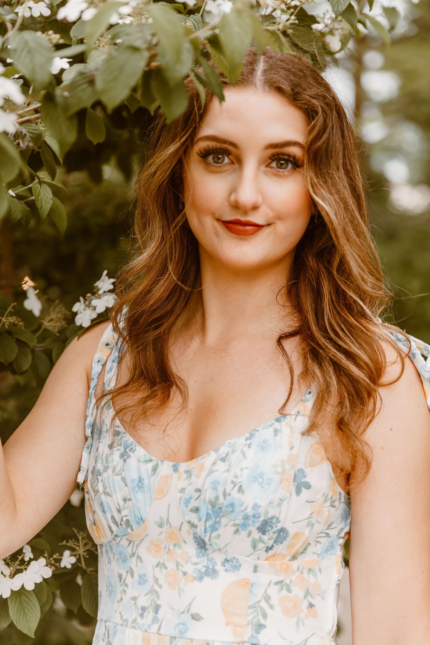 Portrait photograph showing someone in a floral dress standing among spring blossoms in soft natural lighting.