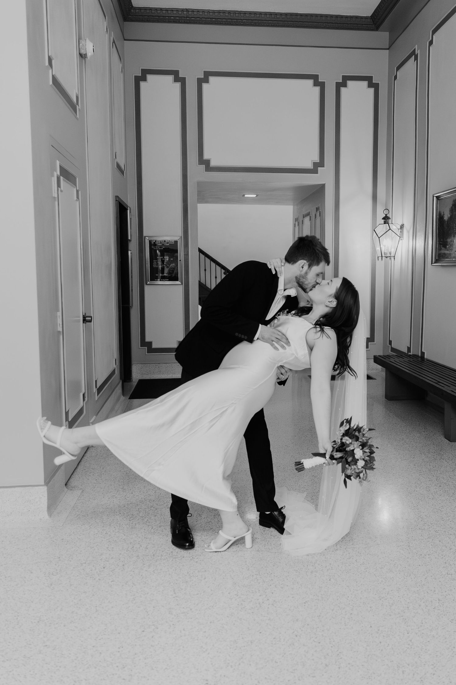 A romantic black and white wedding photo showing a groom dipping his bride in an elegant hallway with decorative molding.