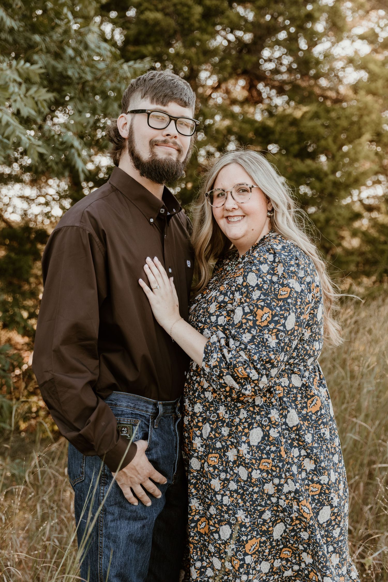 A couple in casual attire poses together outdoors during golden hour in a natural setting.