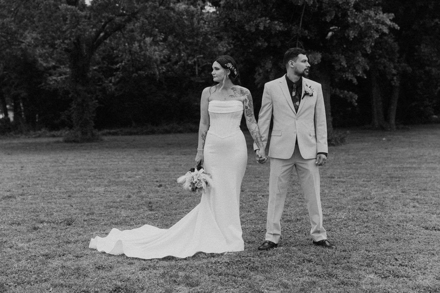 Black and white photo of a bride and groom holding hands outdoors on a grassy lawn surrounded by trees.