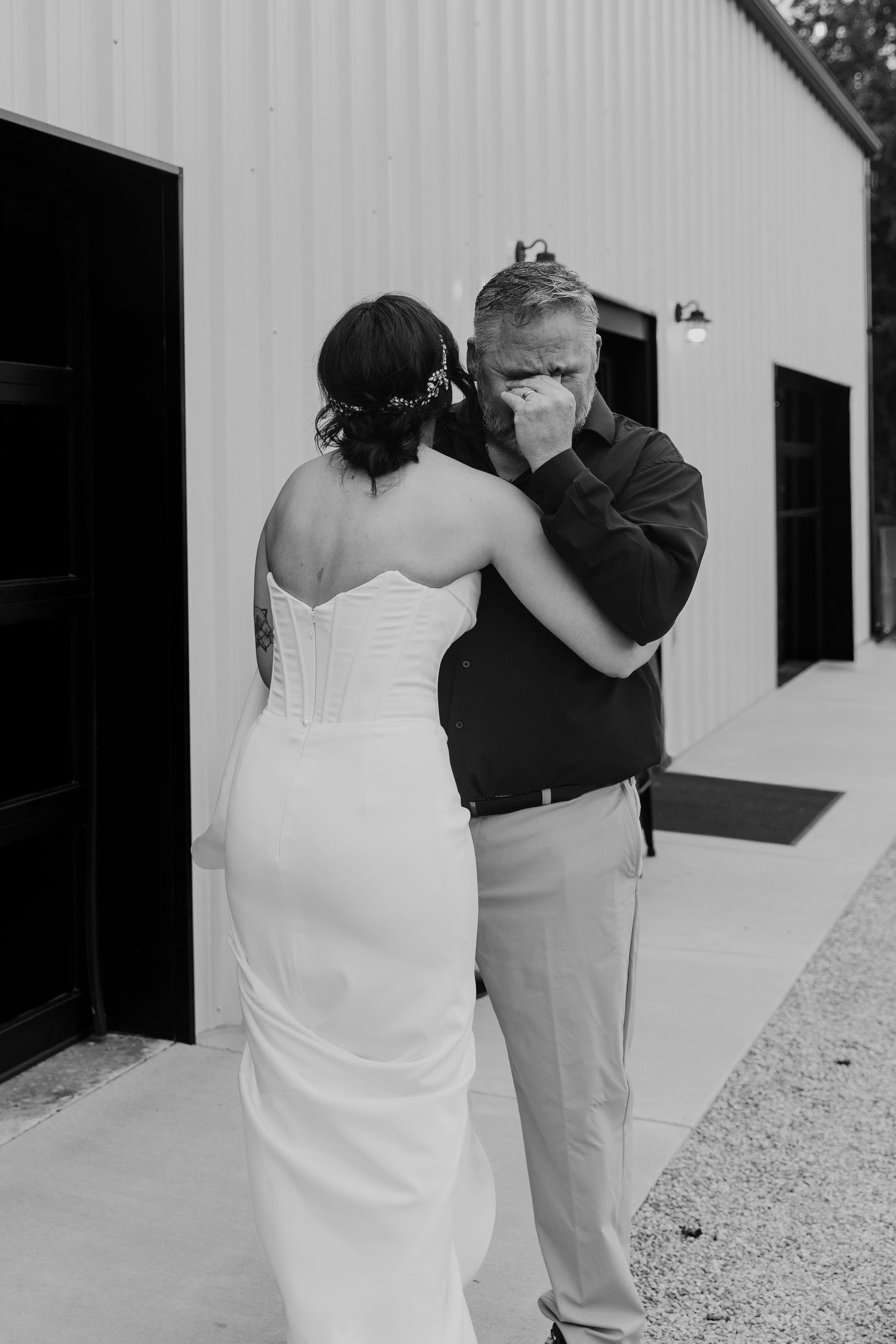 Black and white photo of a bride in a strapless gown sharing an emotional first look moment with a tearful man outside a barn.