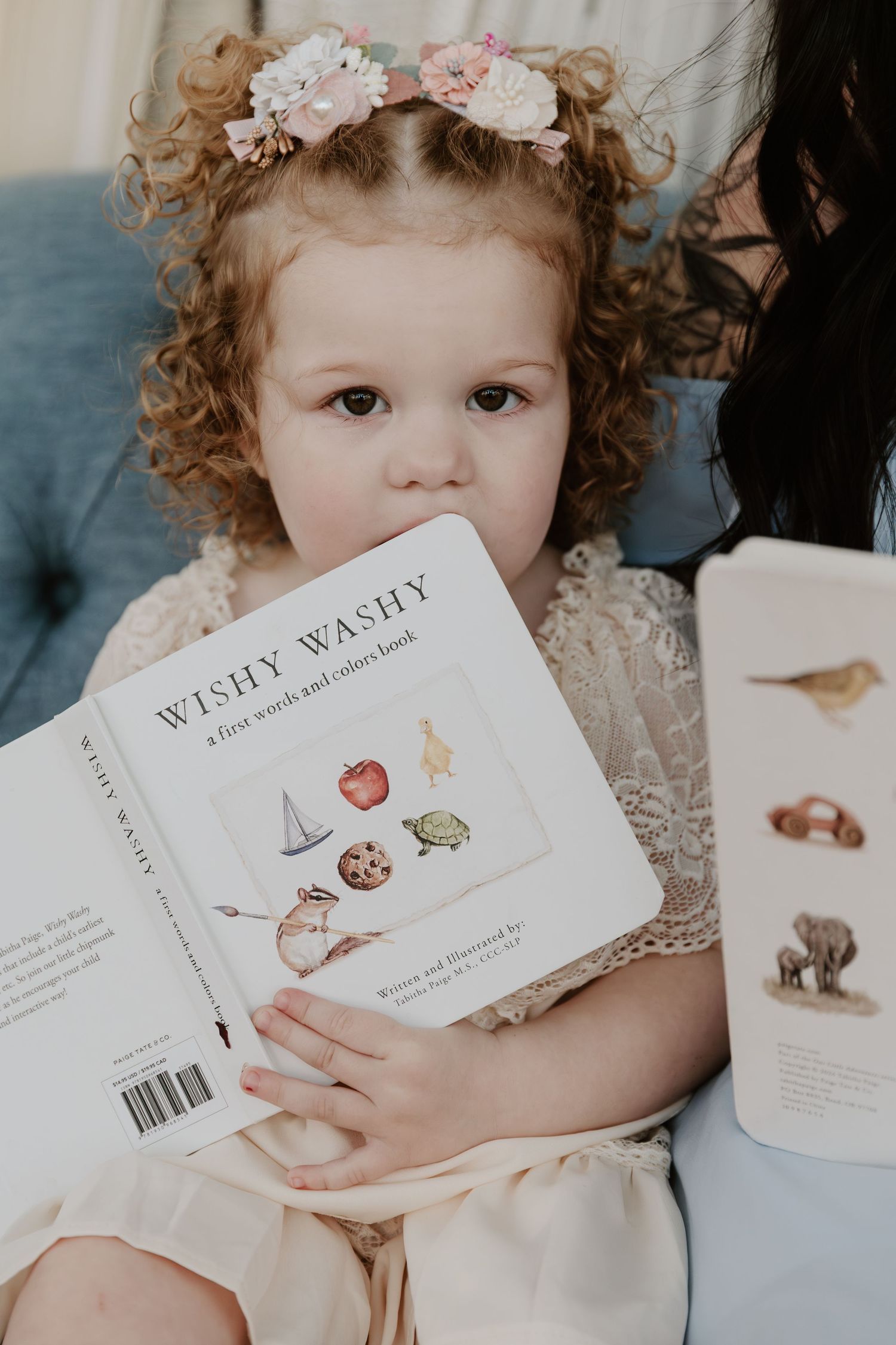A curly-haired toddler girl with floral hair accessories holds the Wishy Washy first words board book.