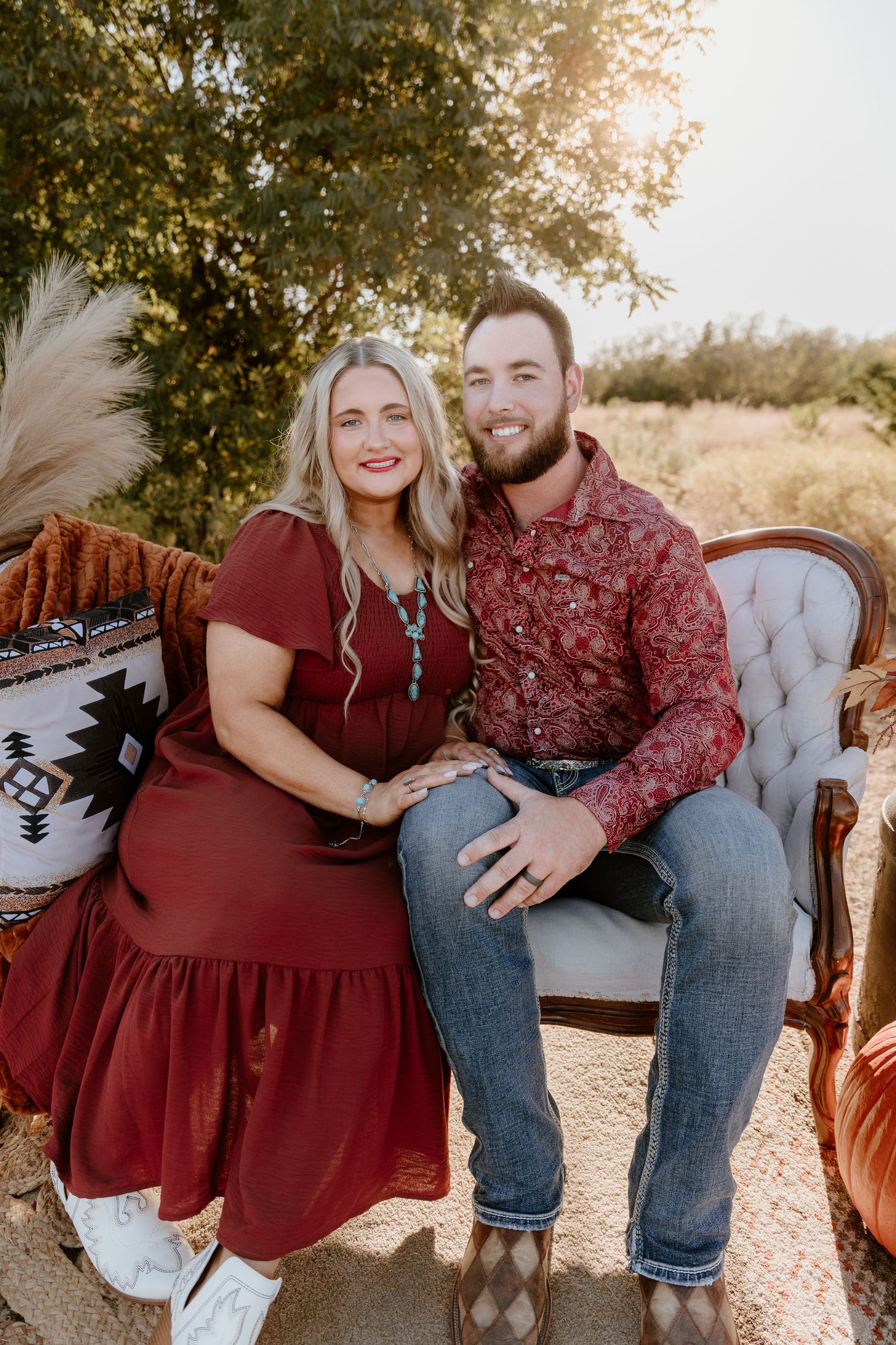 A couple sits together on a vintage sofa outdoors during golden hour wearing burgundy and denim.