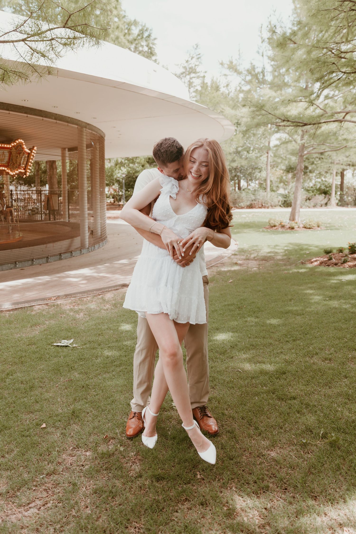 A romantic couple in casual white and tan outfits embrace on a green lawn beside a curved modern building on a sunny day.
