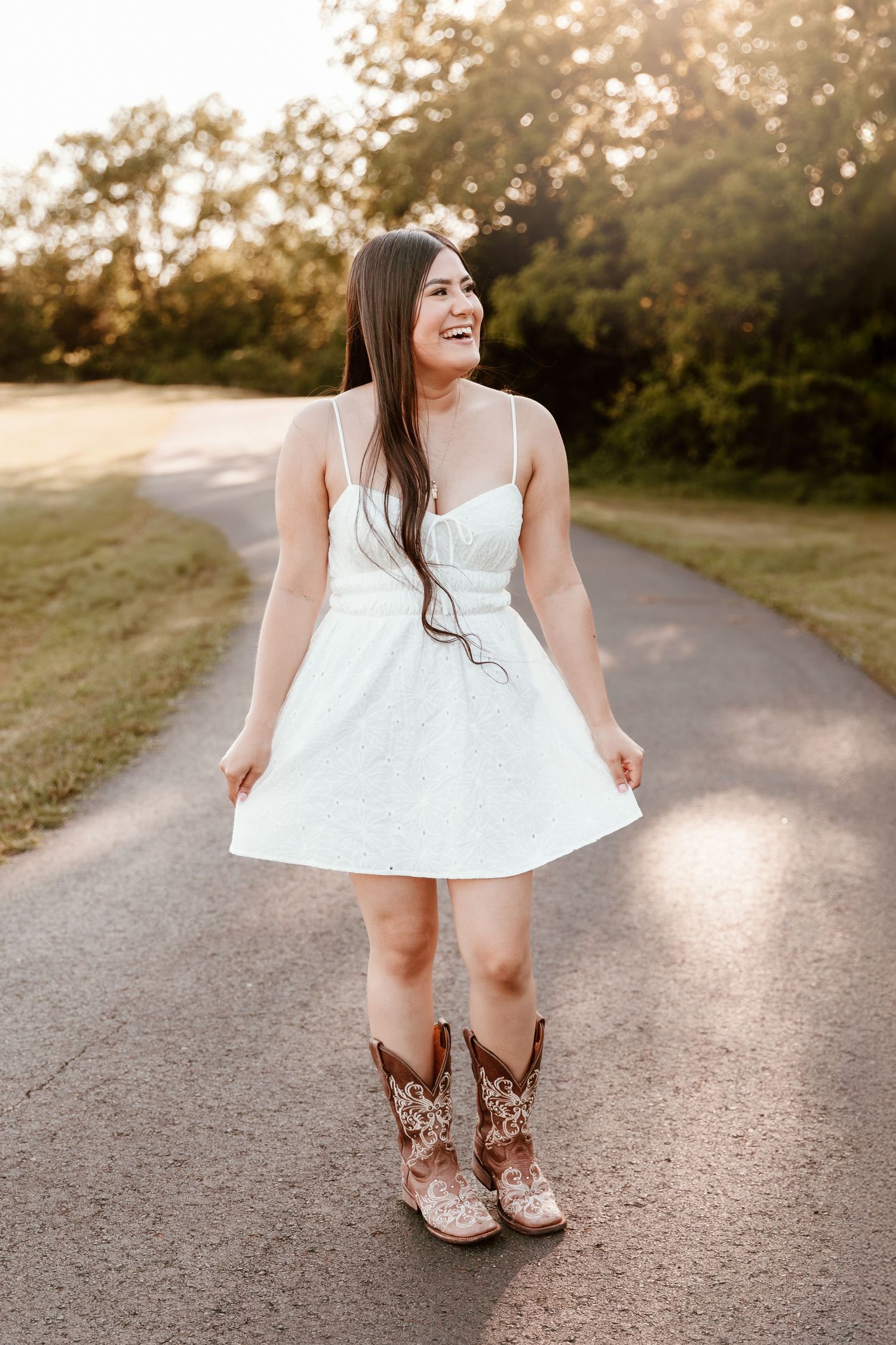 A person in a white summer dress and brown cowboy boots walks along a sunlit gravel path in golden hour lighting.