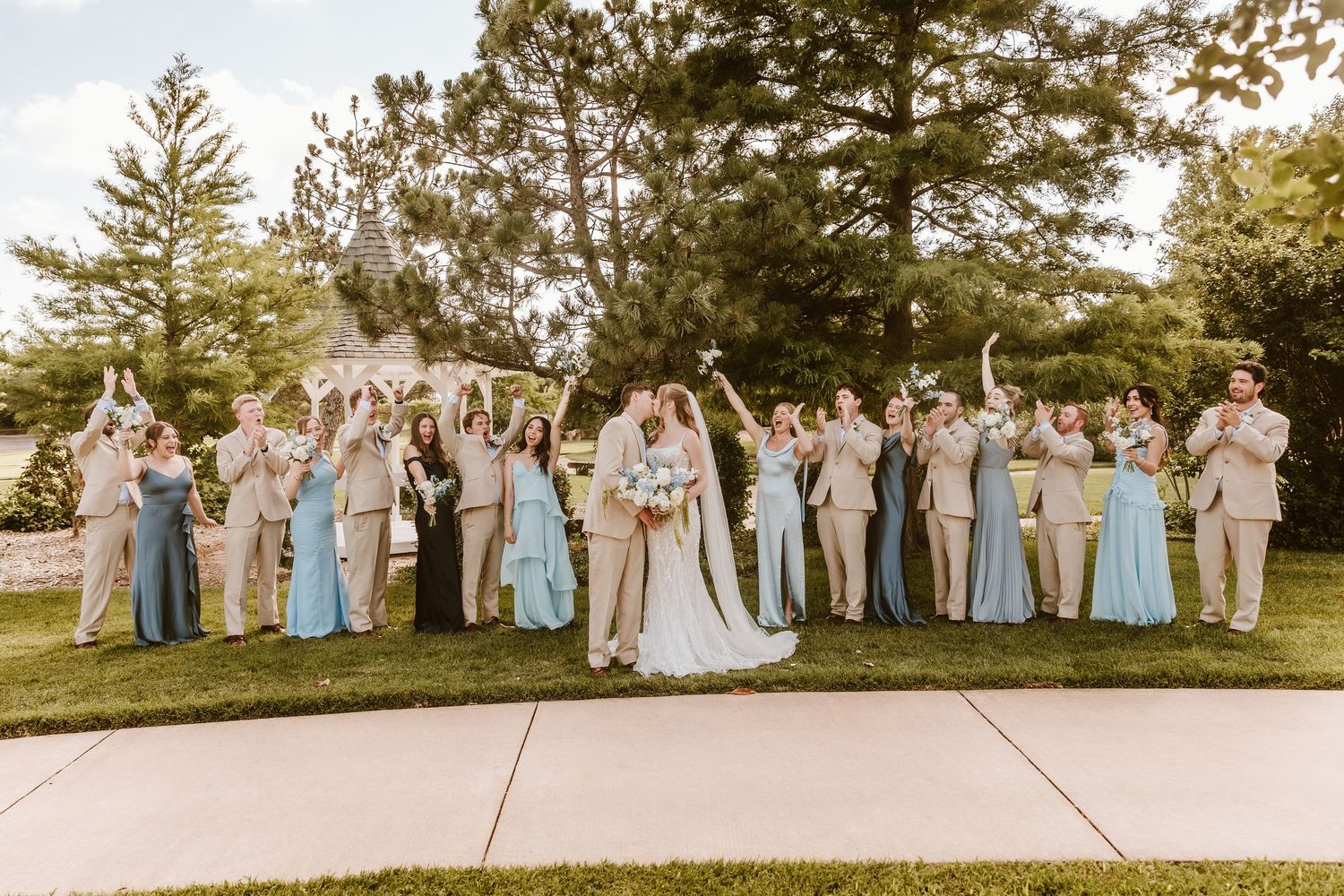 Wedding party celebrating outdoors on green lawn, bride and groom kissing center, bridesmaids in blue gowns, groomsmen in tan suits.