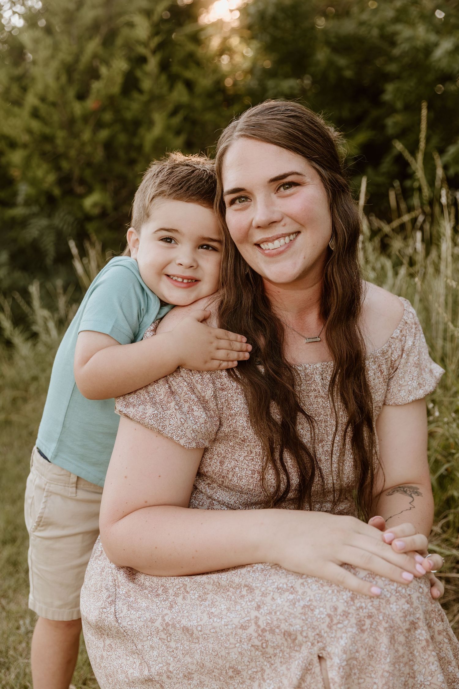 A smiling young woman in a floral dress hugs a happy toddler boy outdoors in a lush green field at golden hour.