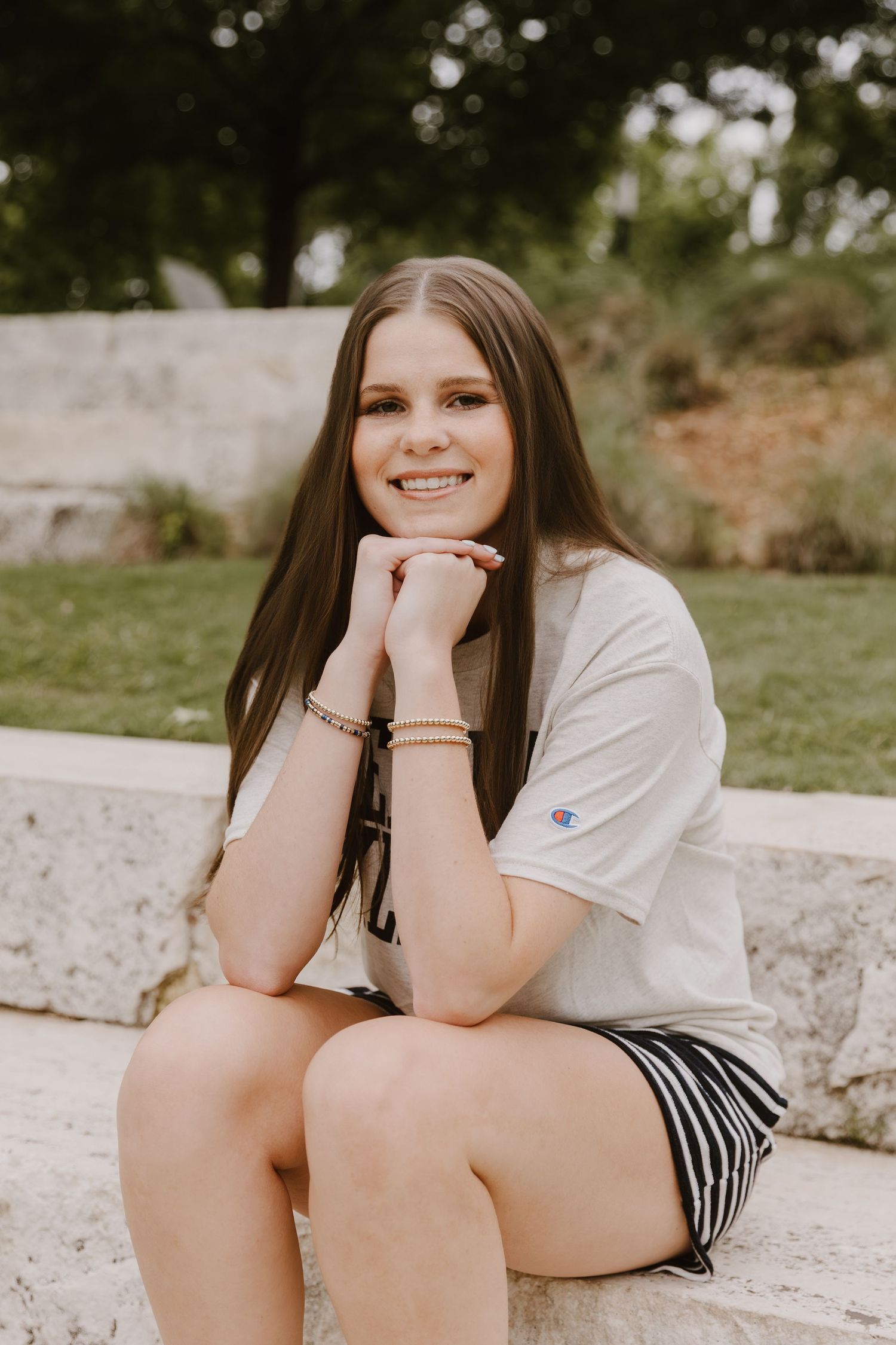 Smiling teenage girl with long brown hair sitting on stone steps outdoors wearing a Champion t-shirt and striped shorts.