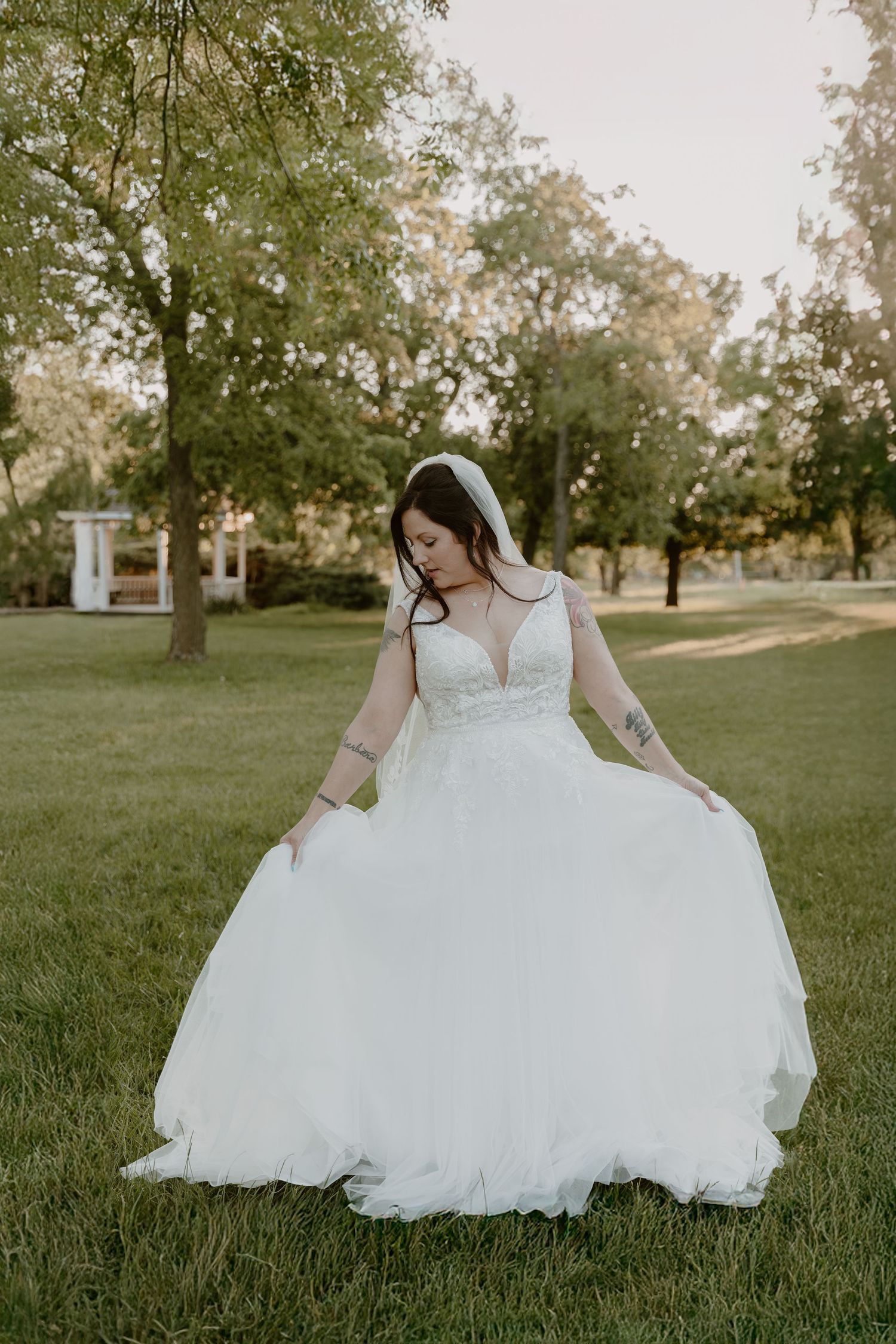 A bride in a flowing white lace and tulle wedding gown poses gracefully on a sunlit lawn surrounded by trees.