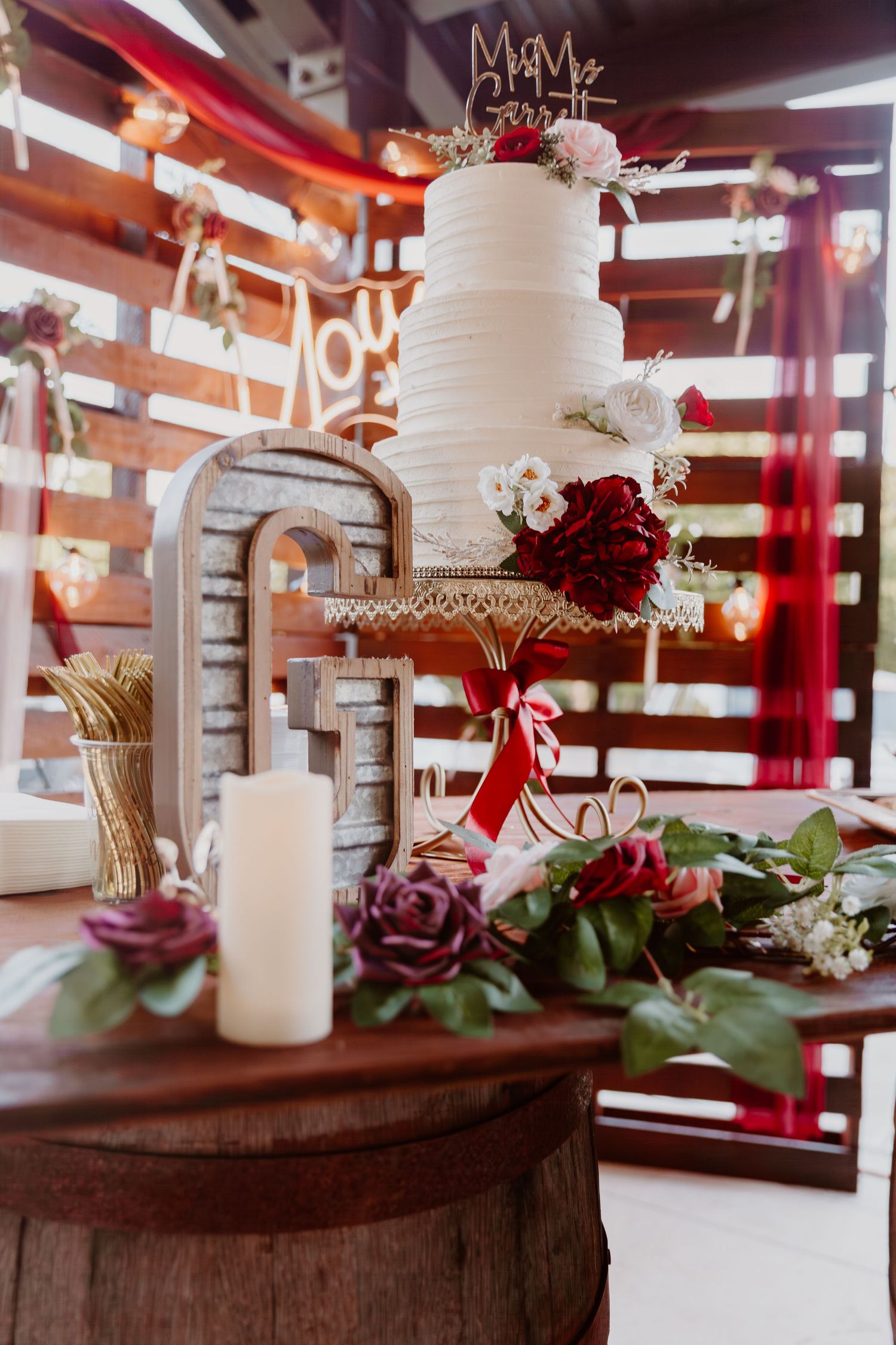 White tiered wedding cake with burgundy floral accents displayed on rustic wooden table with candles and greenery.
