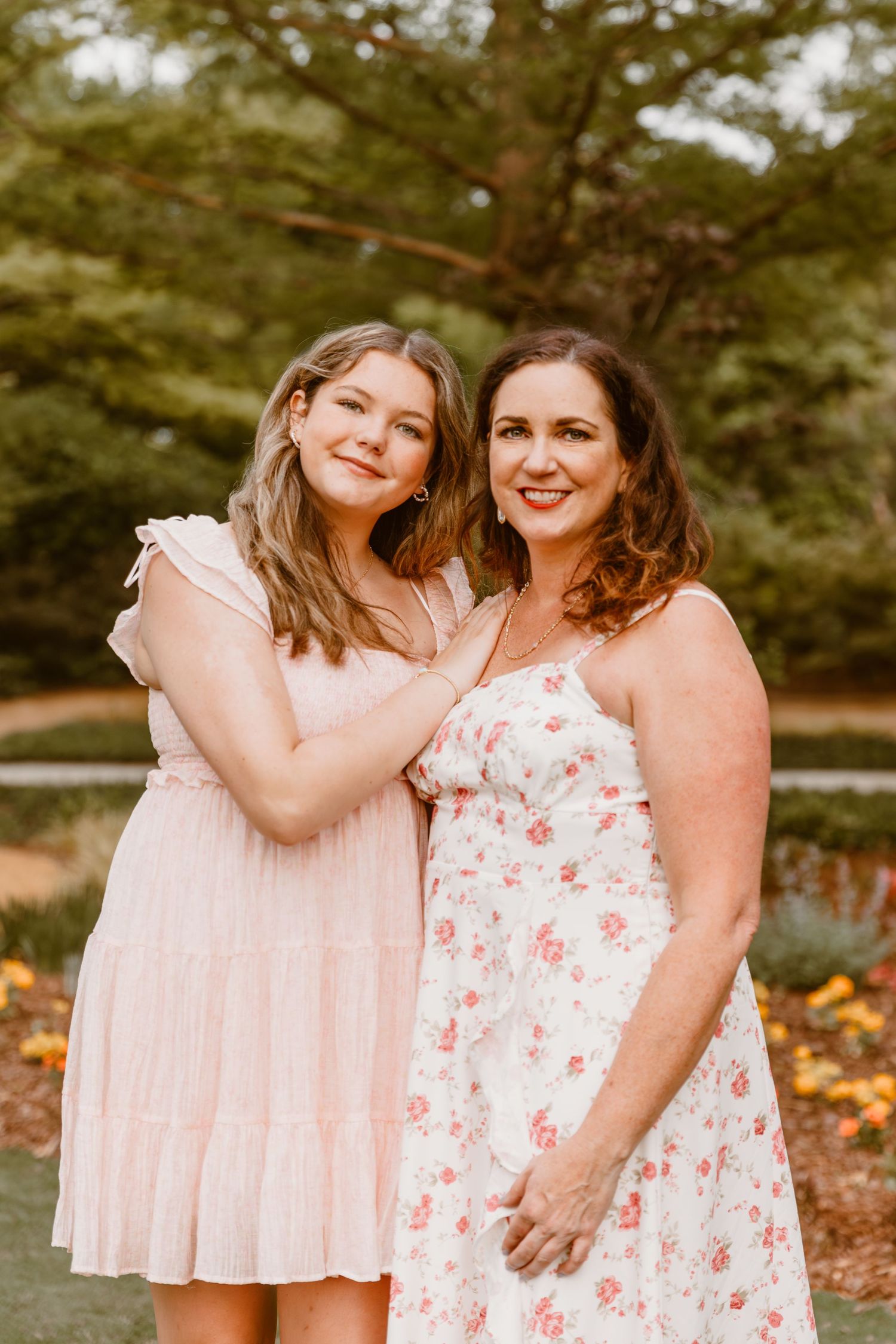 Two women in pink and floral dresses embrace warmly in a natural outdoor autumn setting.