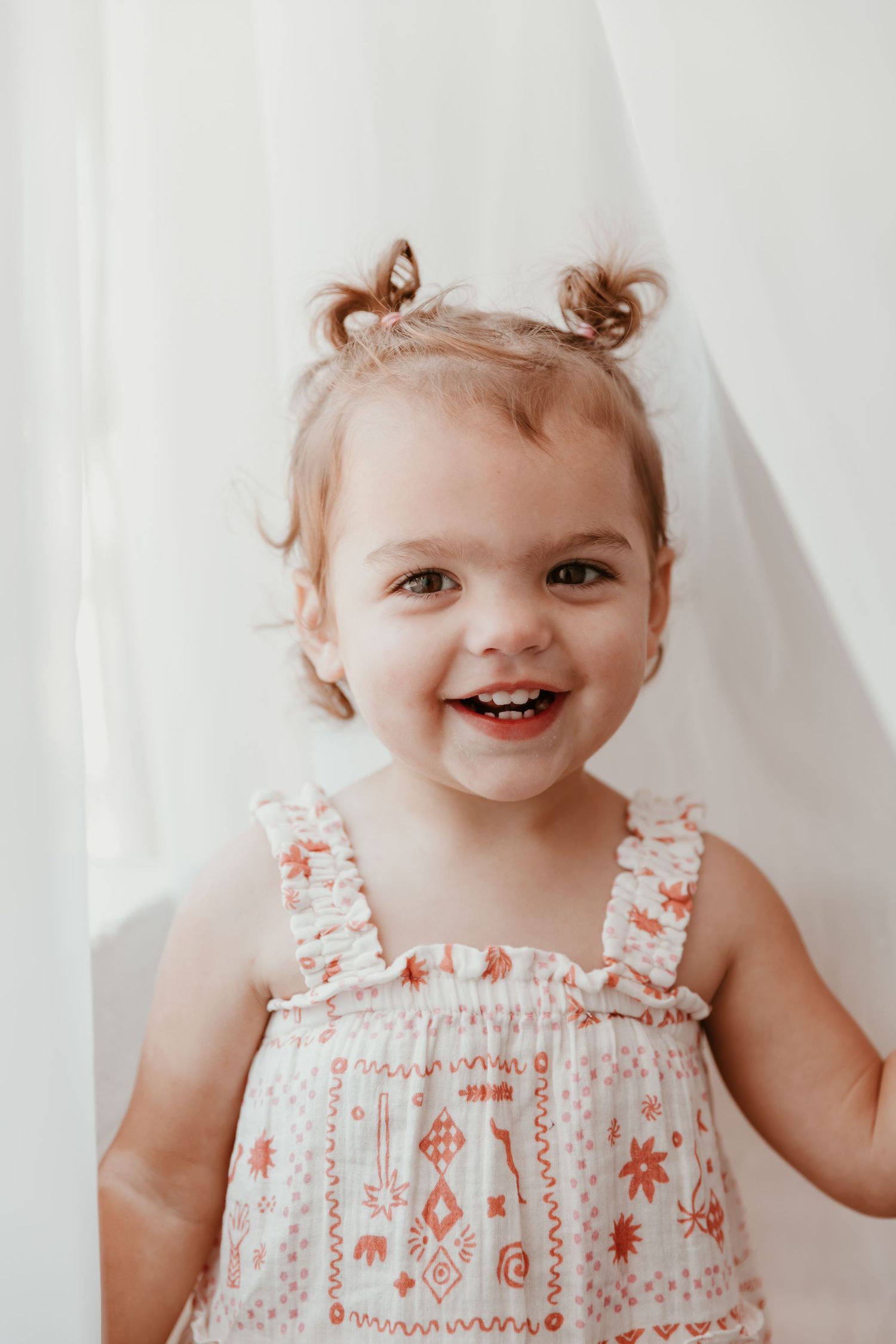 Smiling toddler wearing a white and pink patterned sundress with small pigtails against a light background.